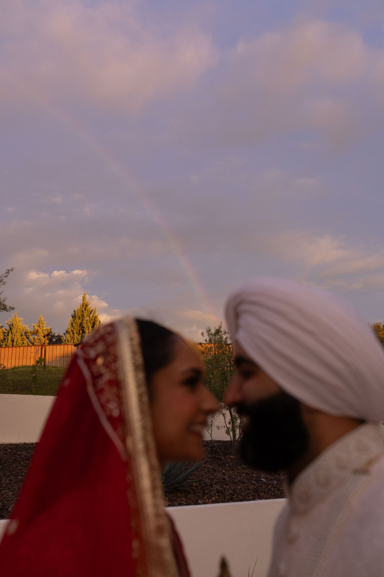 A romantic moment captured at sunset with a beautiful rainbow in the background during a traditional Sikh wedding ceremony.