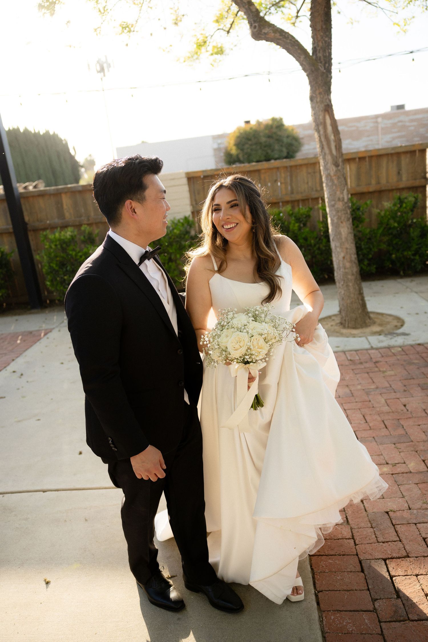 A bride in a white gown holding a bouquet smiles at her groom in a black tuxedo on a brick patio.