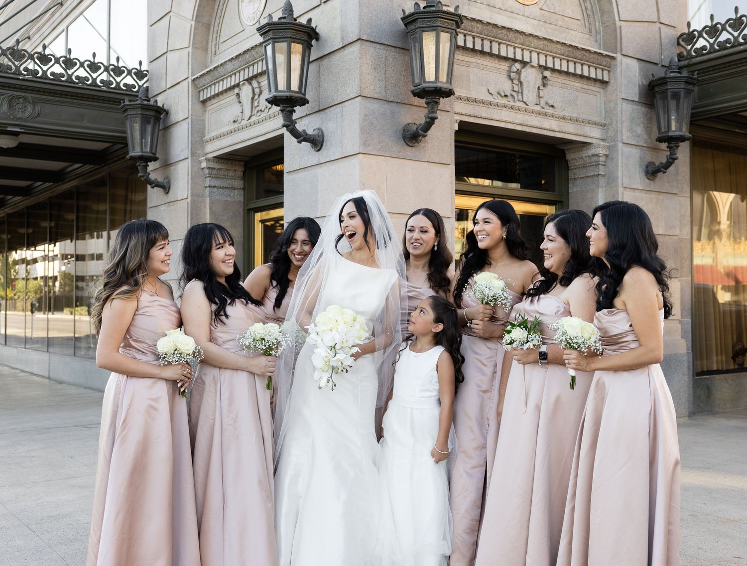 Wedding party in blush pink bridesmaid dresses gather around bride outside classic stone building.