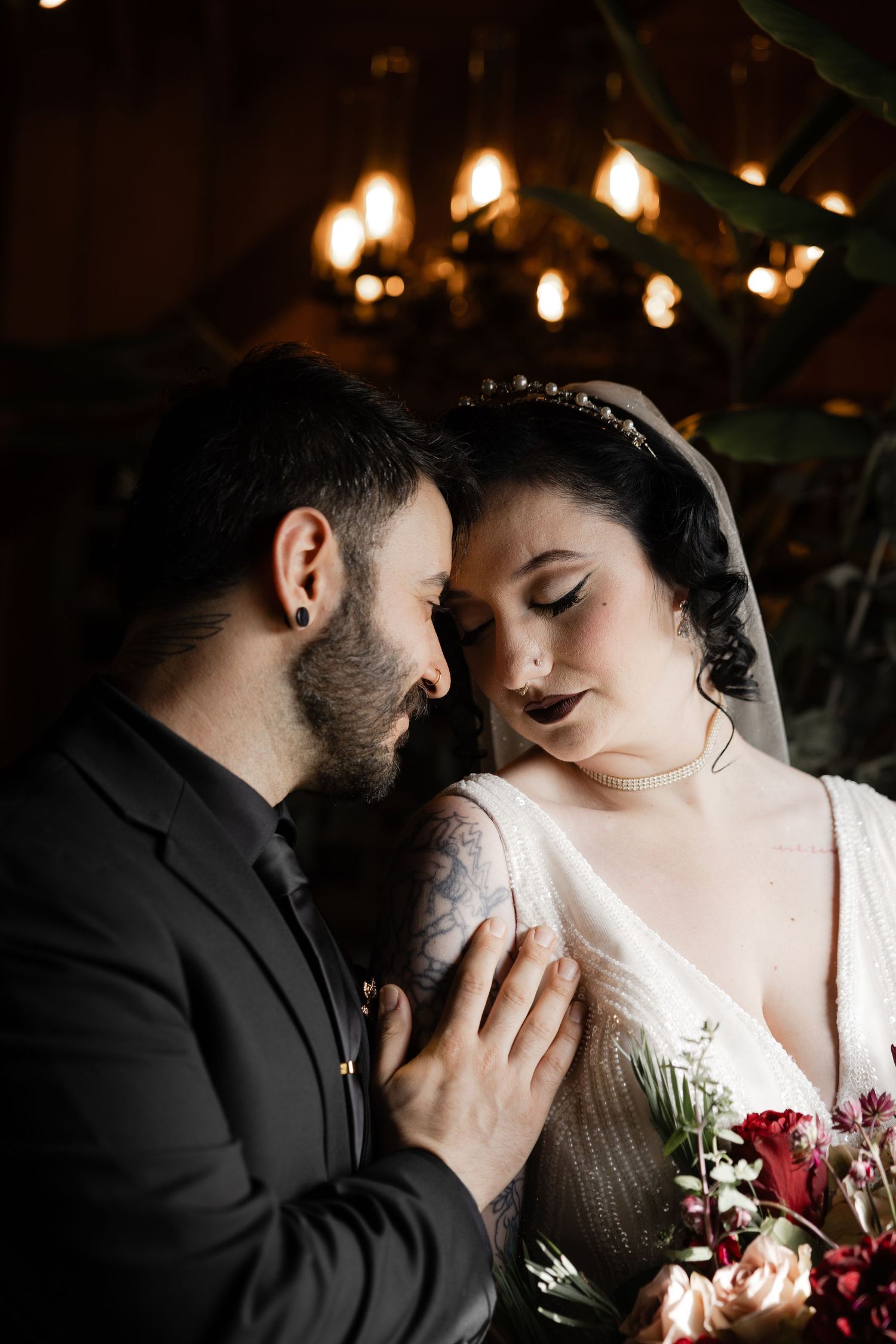 Romantic wedding couple shares an intimate moment with soft lighting and red floral bouquet details.
