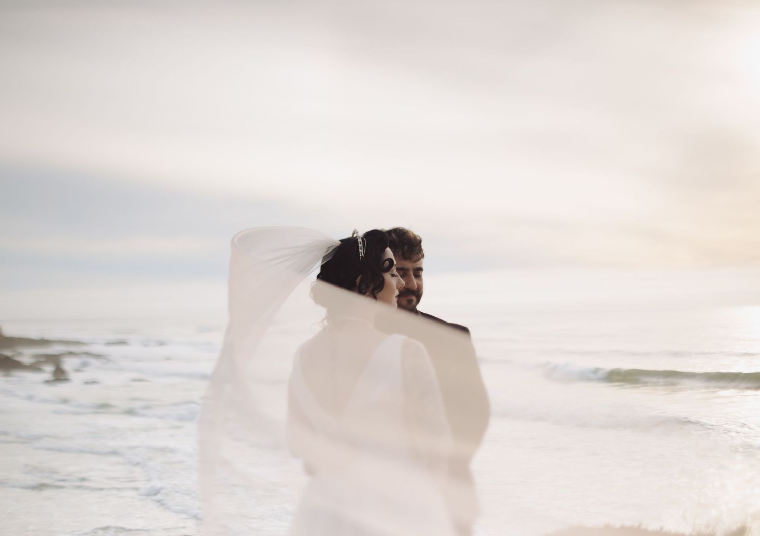 Two silhouettes stand on a coastal cliff overlooking the ocean at sunset while white fabric billows in the wind.