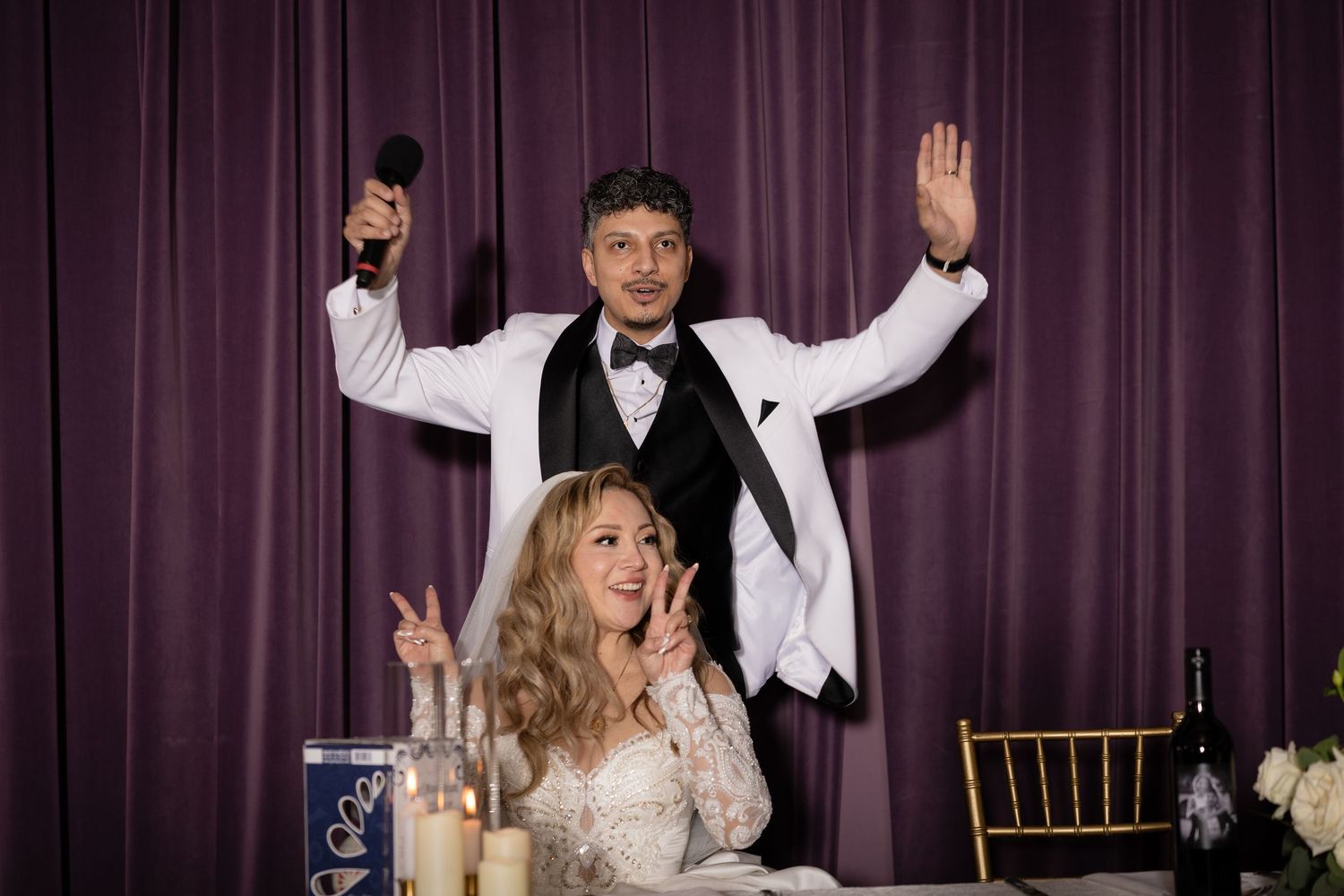 A celebratory moment at a wedding reception with raised hands against a purple curtain backdrop.