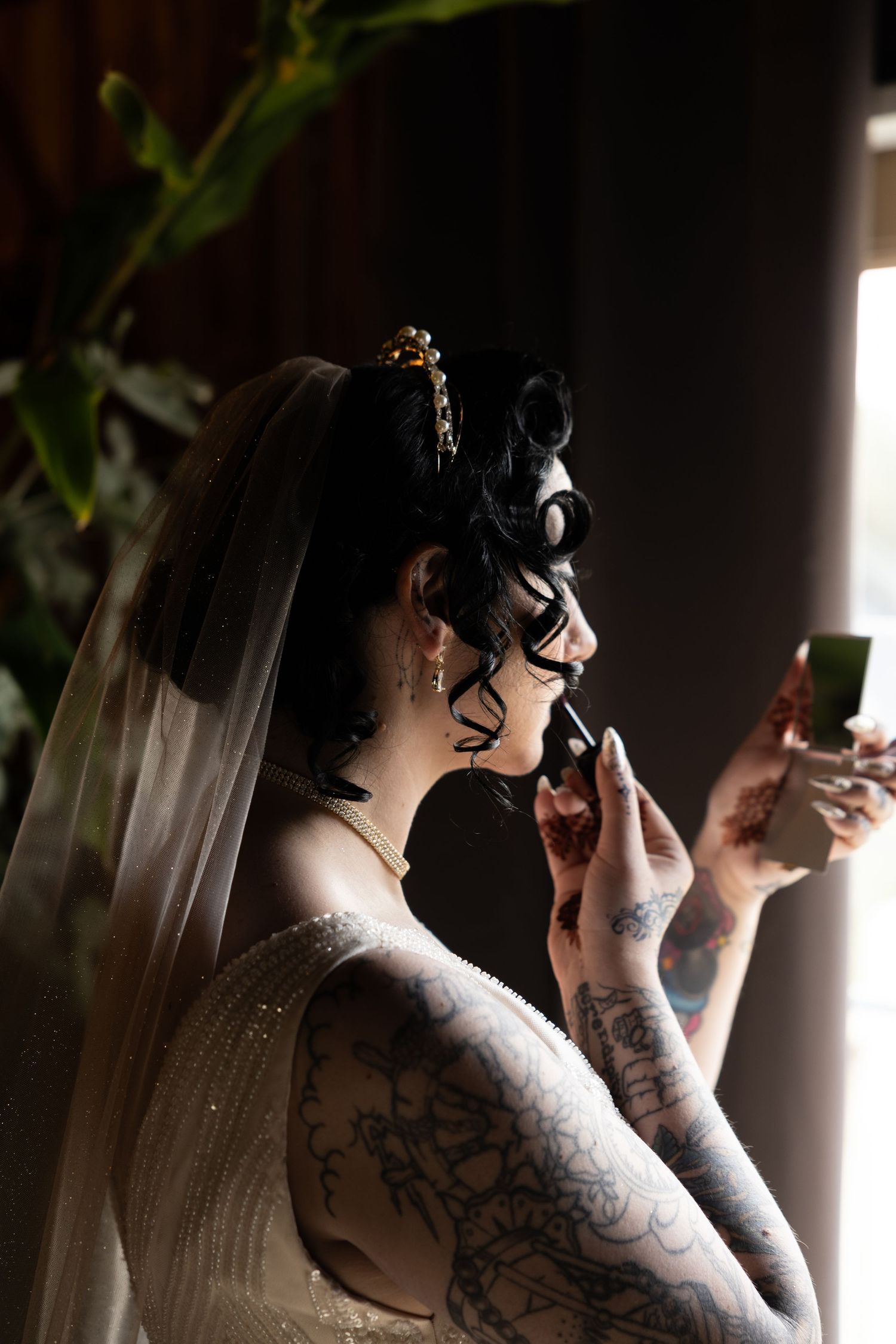 A bride with tattoos applies makeup while wearing a white dress and veil in dramatic lighting.
