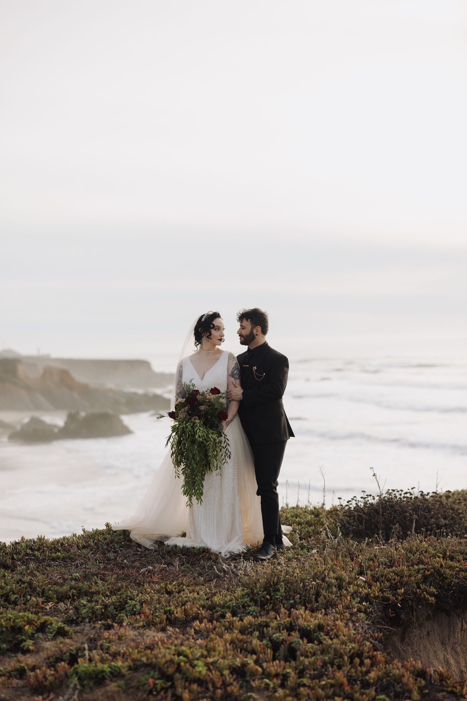 A romantic wedding portrait on coastal cliffs with dramatic ocean views and overcast skies.