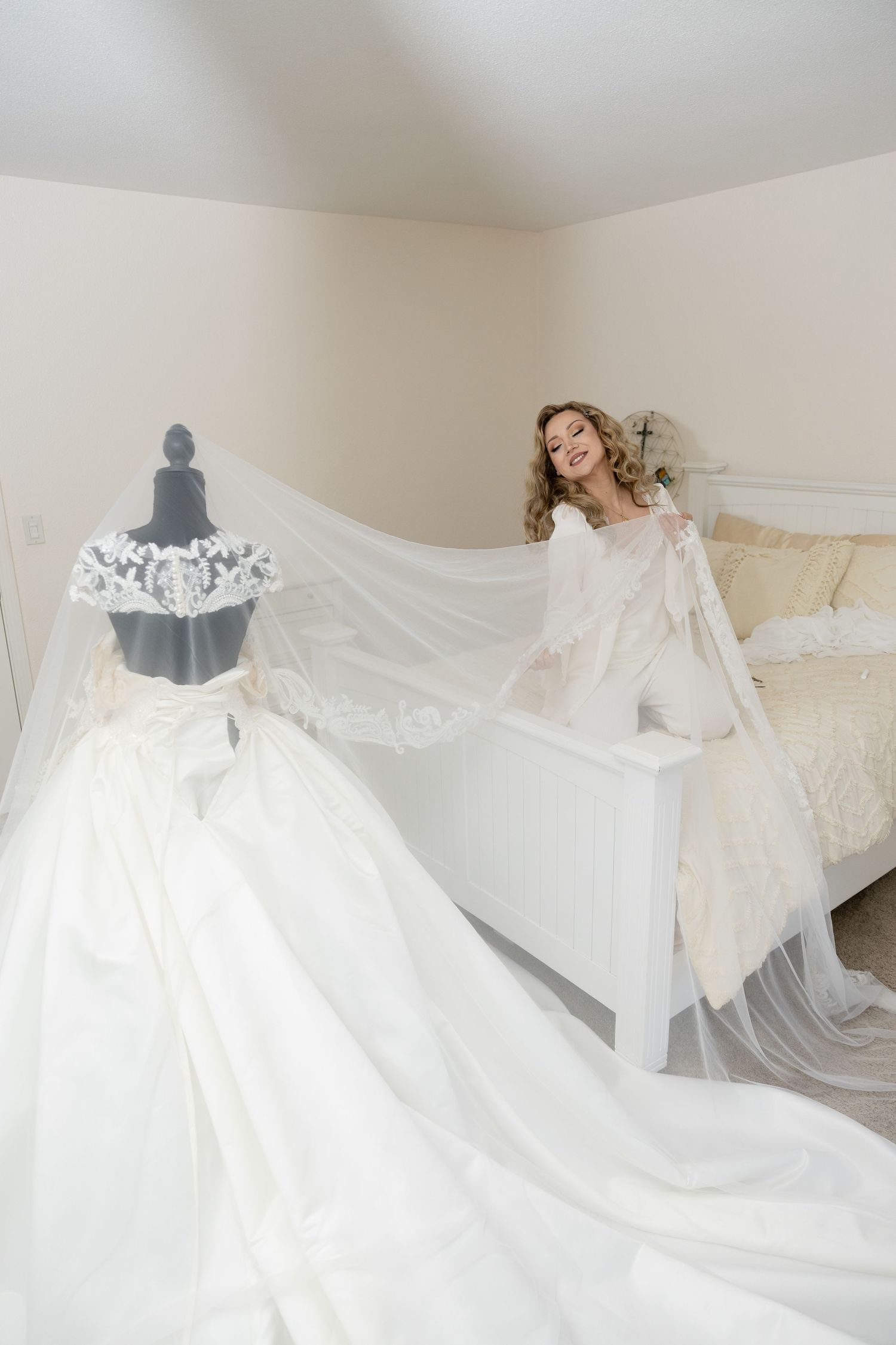 Wedding dress displayed on a mannequin with flowing veil in a bright bedroom setting.