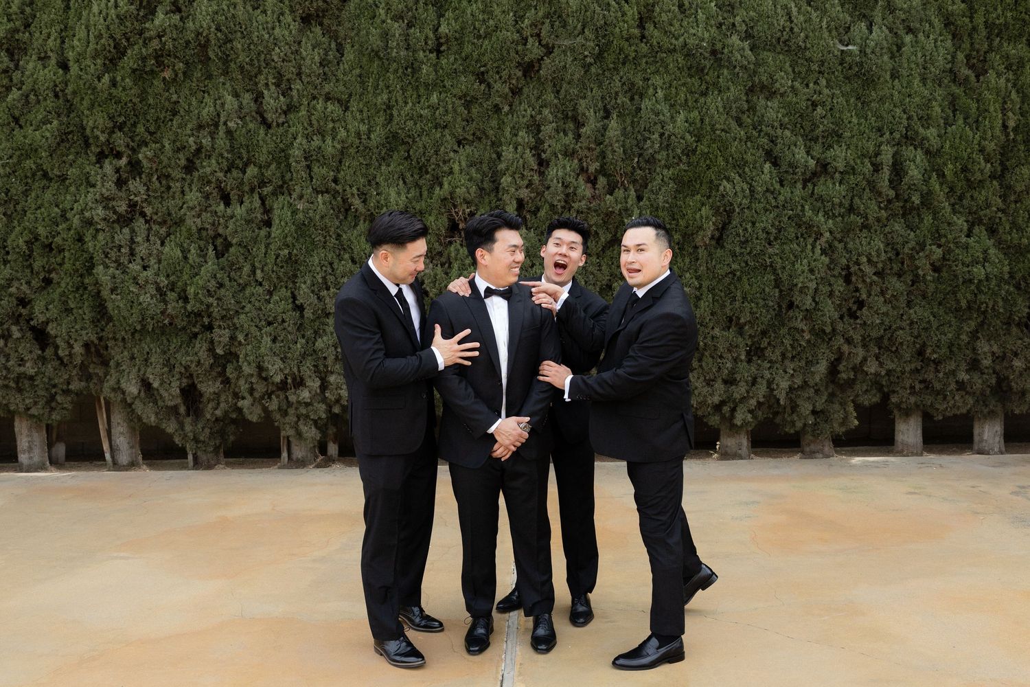 Four groomsmen in black tuxedos laughing and celebrating together outdoors before a wedding ceremony.