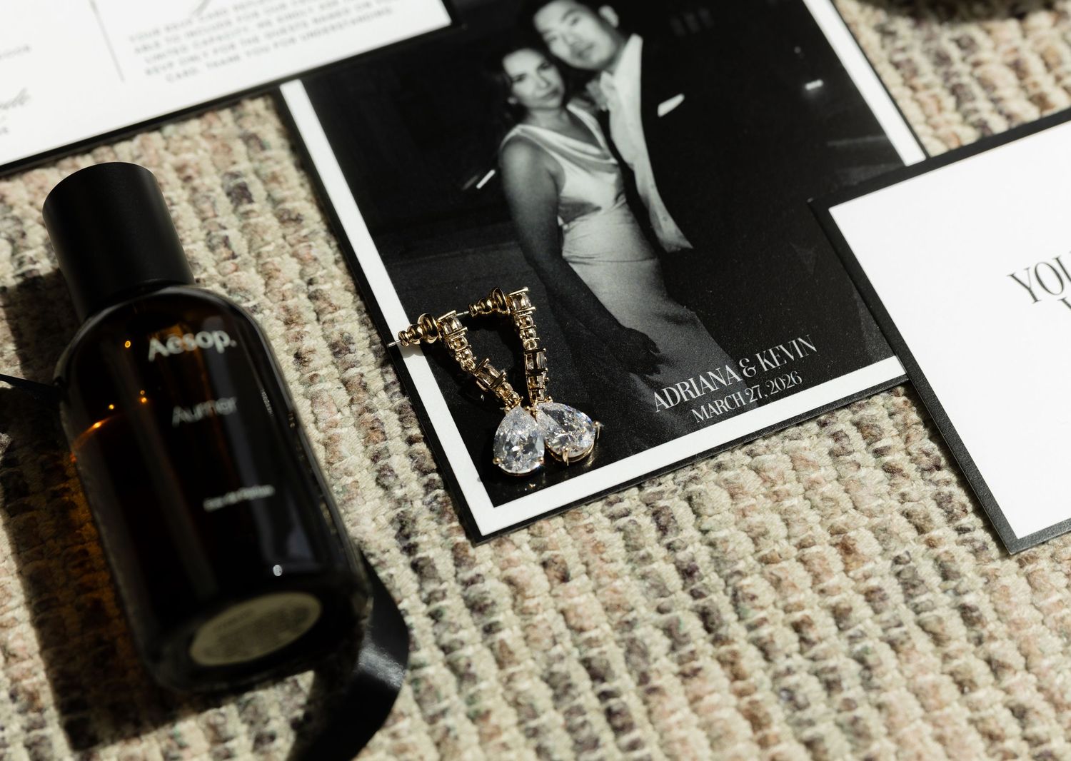 Aesop perfume bottle beside a gold crystal necklace and black-and-white wedding photo card on a woven rug.