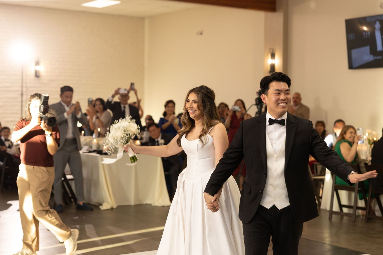 Bride and groom smile while making grand entrance at elegant wedding reception hall filled with seated guests.