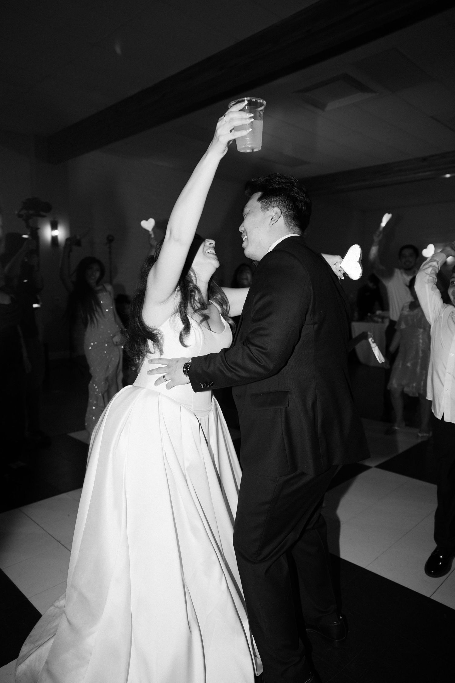 Black and white photo of bride and groom dancing joyfully, bride raising a drink at wedding reception.