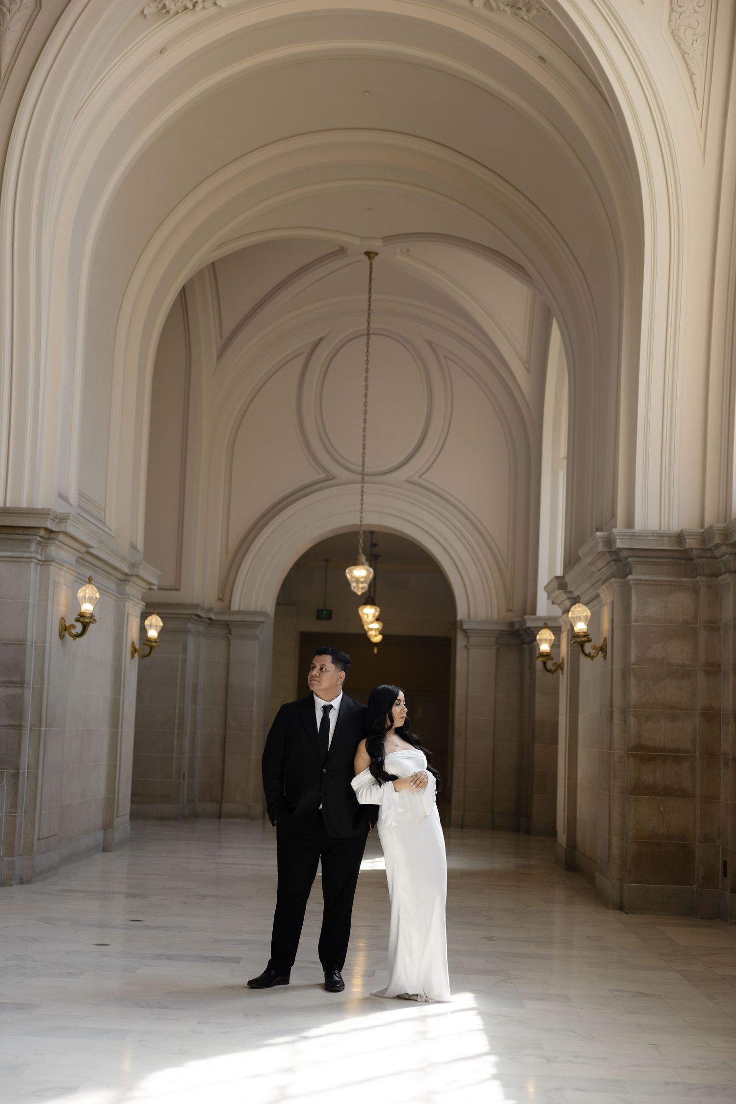 A couple stands in formal attire under arched ceilings with pendant lights in a classical corridor.