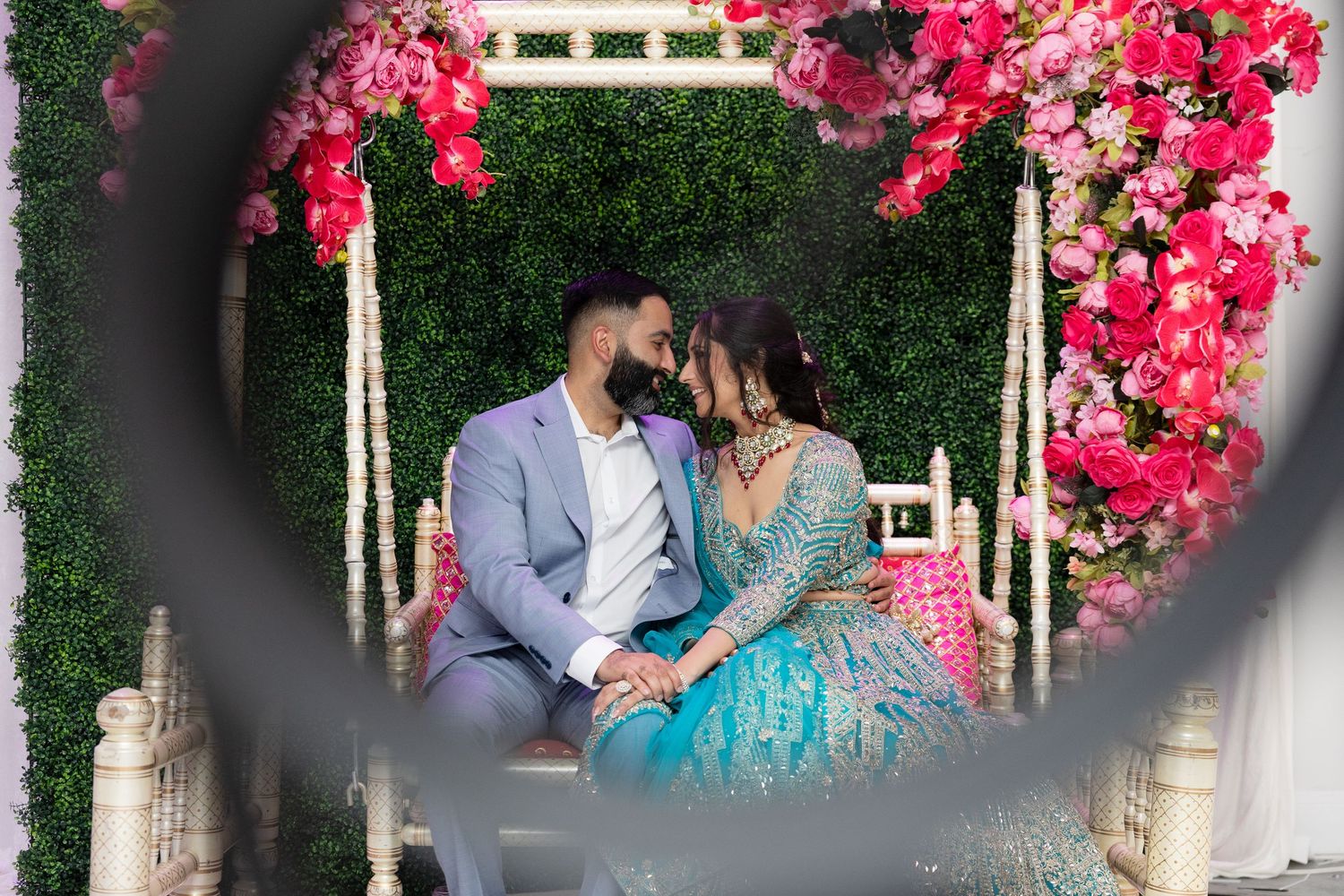 A romantic couple sits on a decorated swing adorned with vibrant pink flowers and greenery at an outdoor celebration.