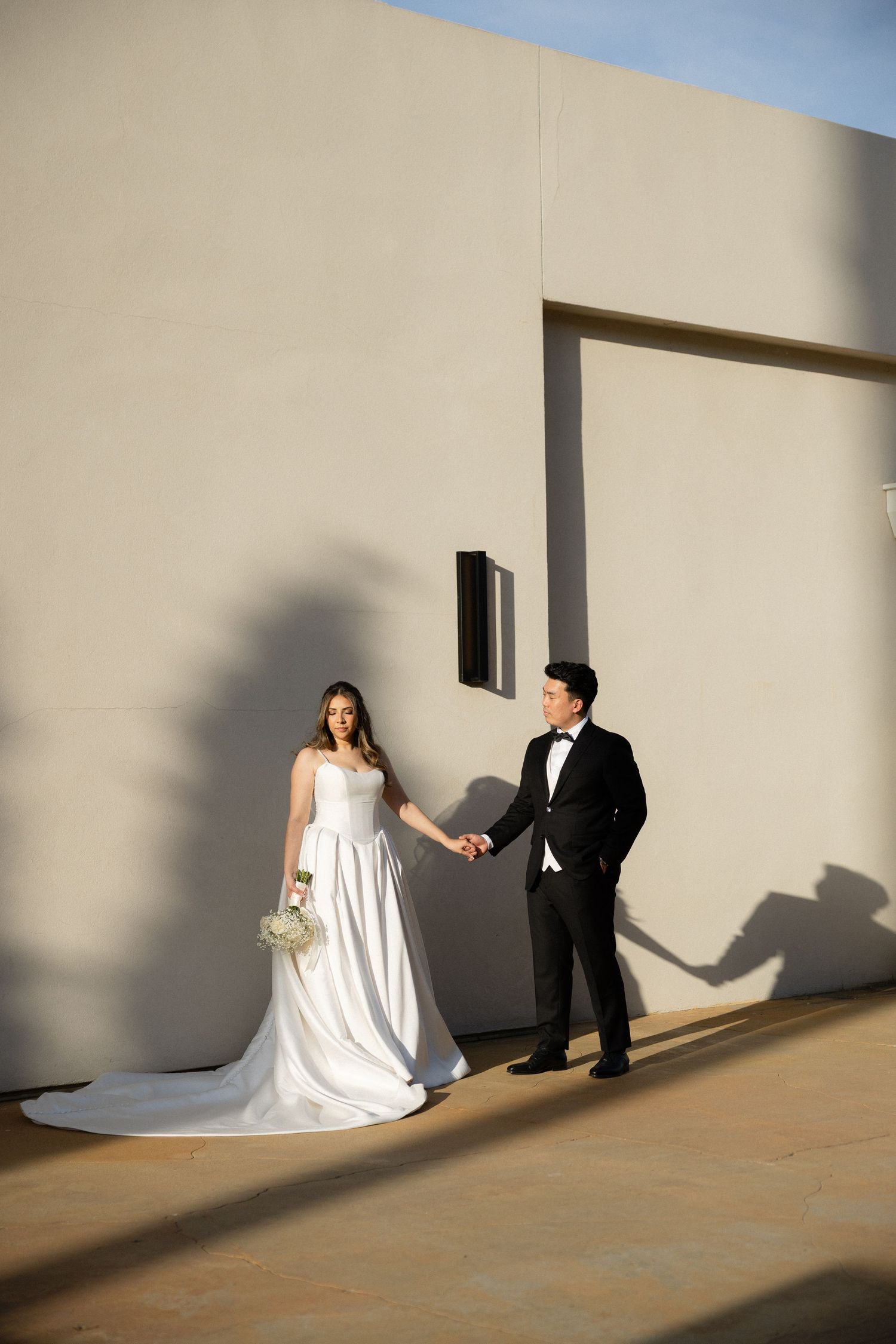 Bride in white gown and groom in tuxedo hold hands against a sunlit beige wall, casting romantic shadows.