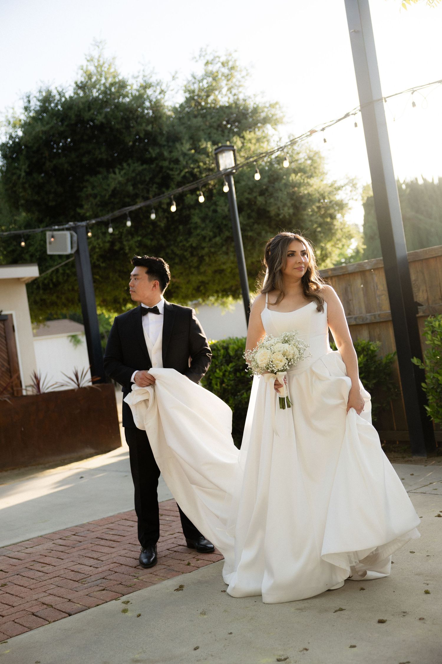 Bride in white gown holding bouquet walks with groom in tuxedo carrying her train at outdoor wedding venue.