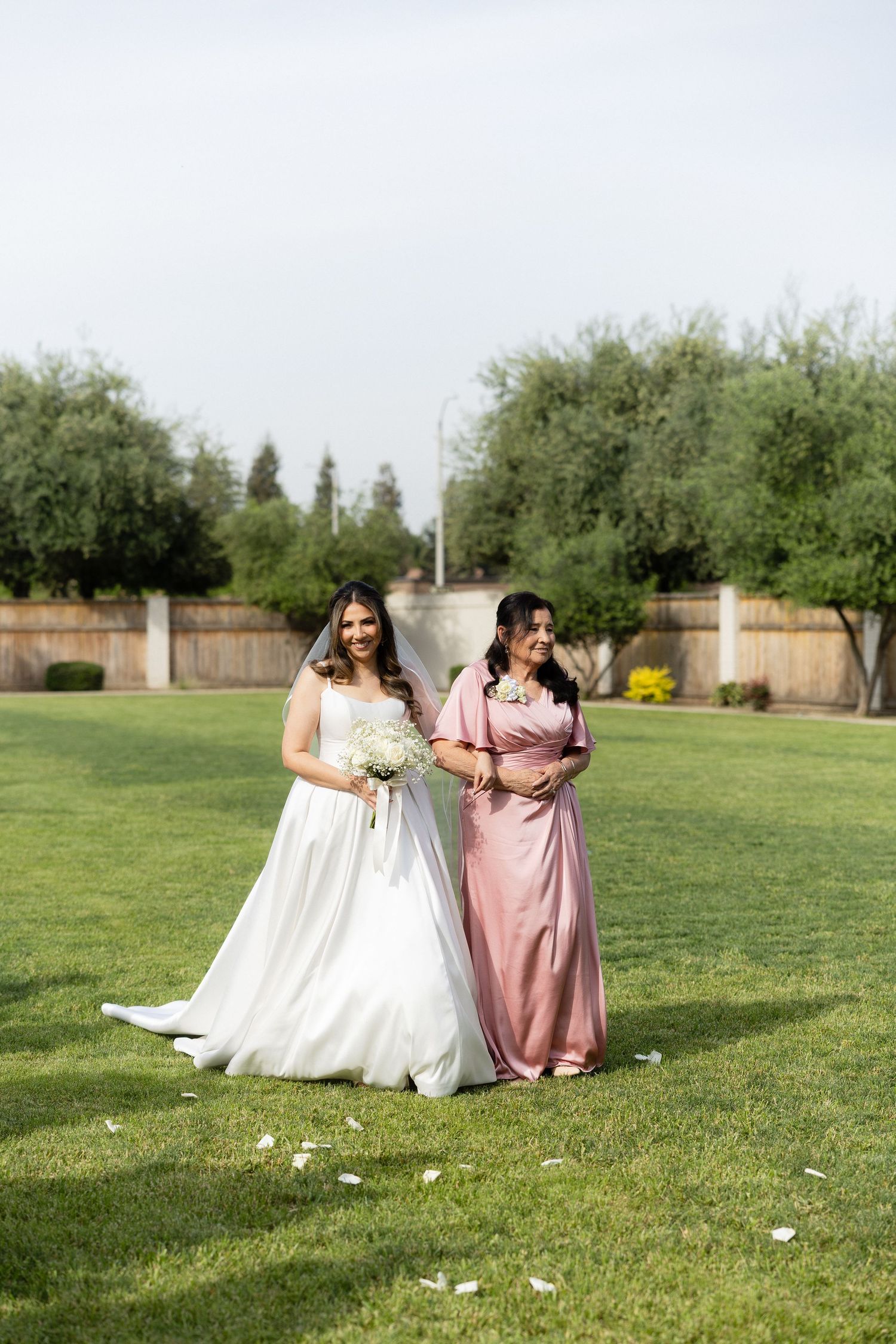 Bride in white gown with veil walks with woman in pink dress across green lawn at wedding.