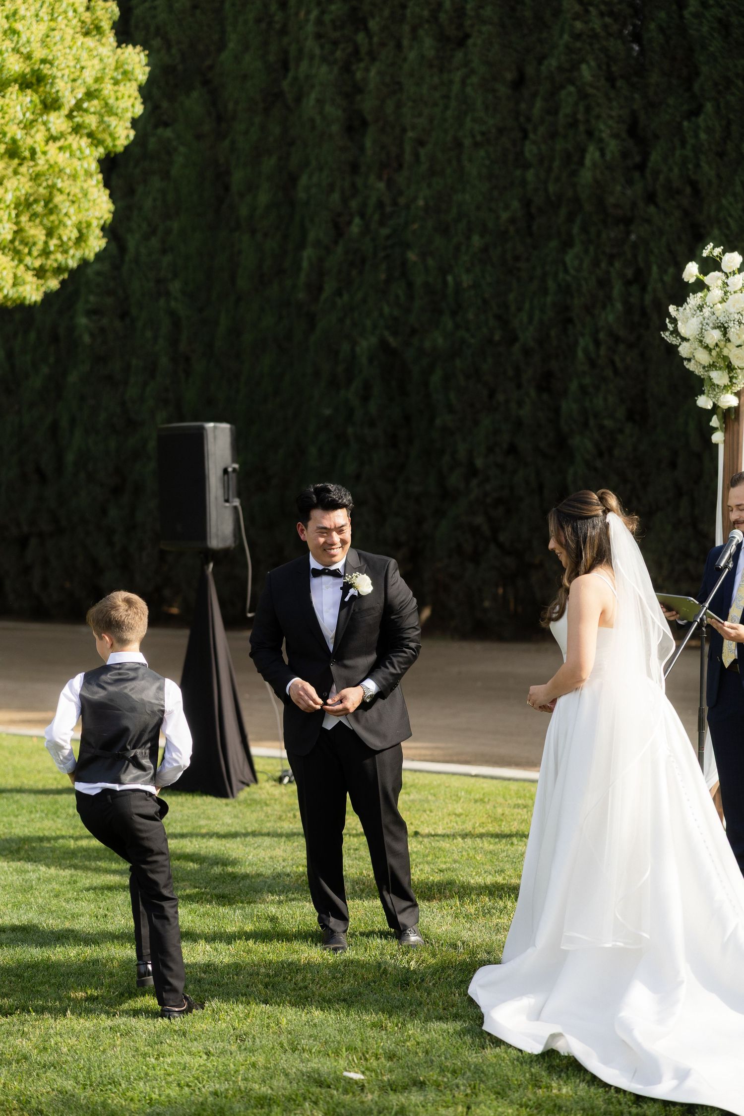 Groom in black tuxedo smiling at bride in white gown during outdoor wedding ceremony on green lawn.
