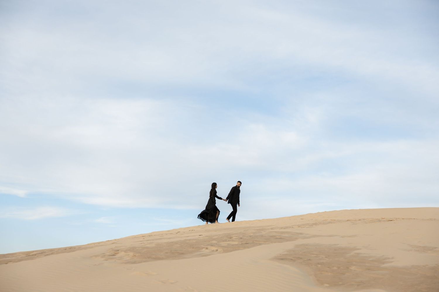 Two silhouettes stand atop a sweeping sand dune against a cloudy sky at sunset.