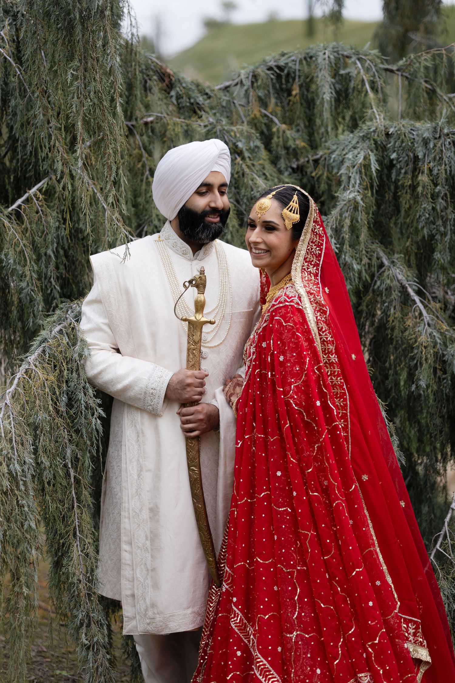 A couple in traditional Sikh wedding attire poses together under willow trees.