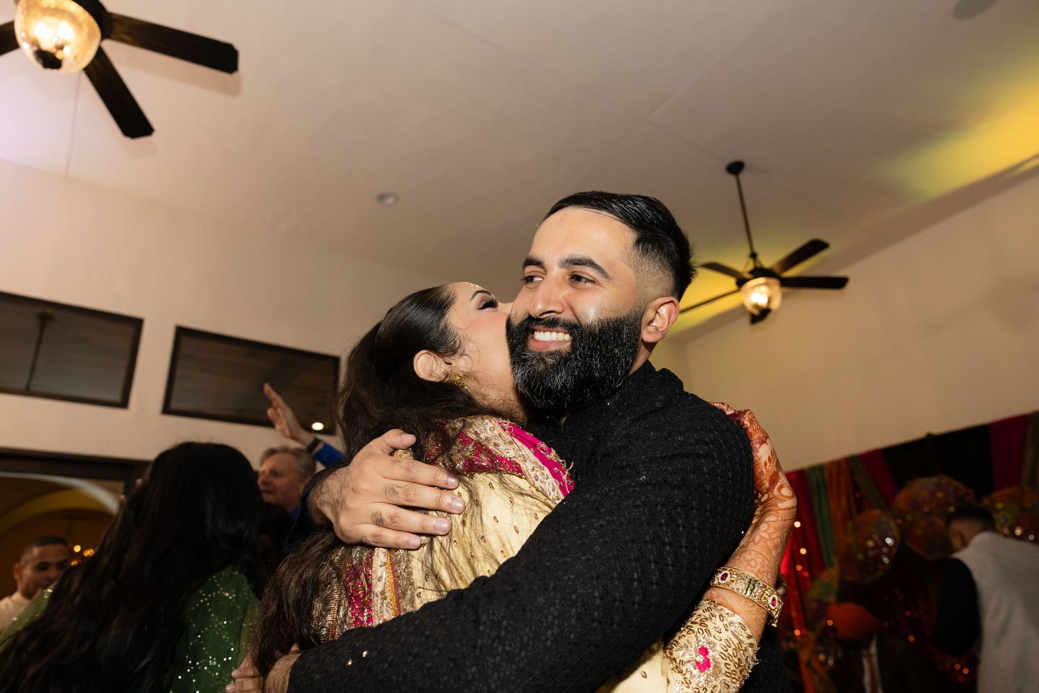 Couple sharing an intimate embrace at an indoor wedding reception with ceiling fans visible.