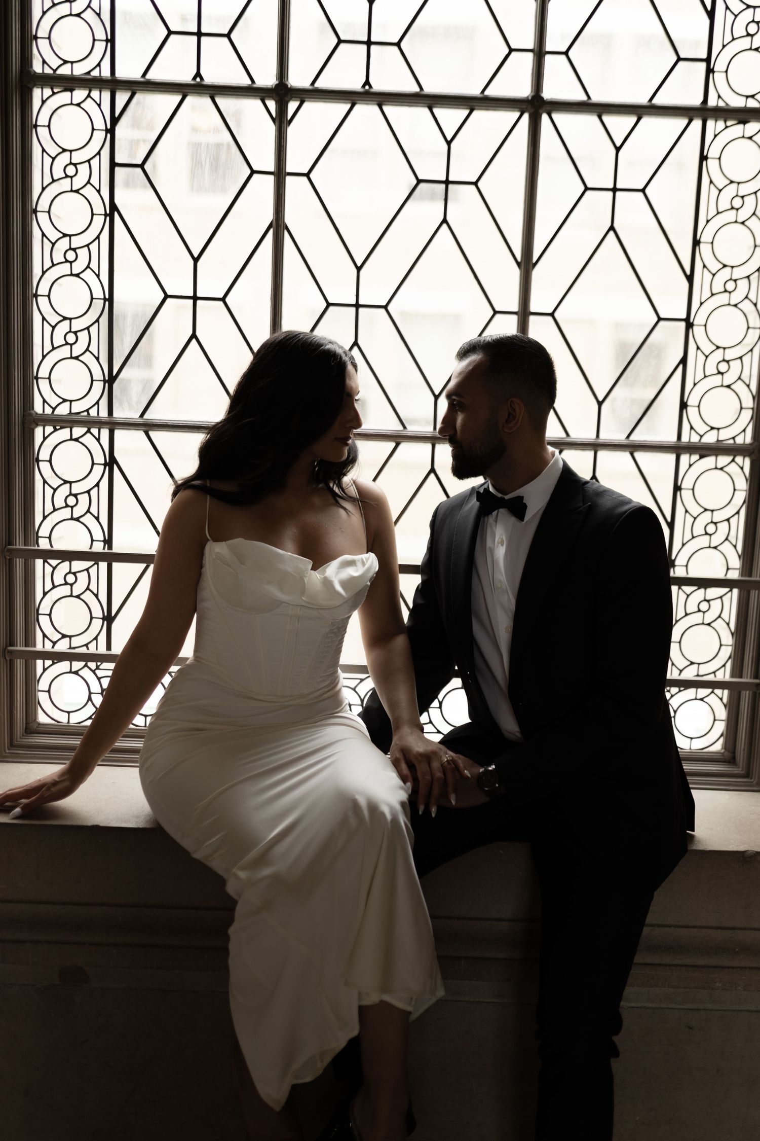 A romantic silhouette of a couple in wedding attire sitting by an ornate window with geometric patterns.
