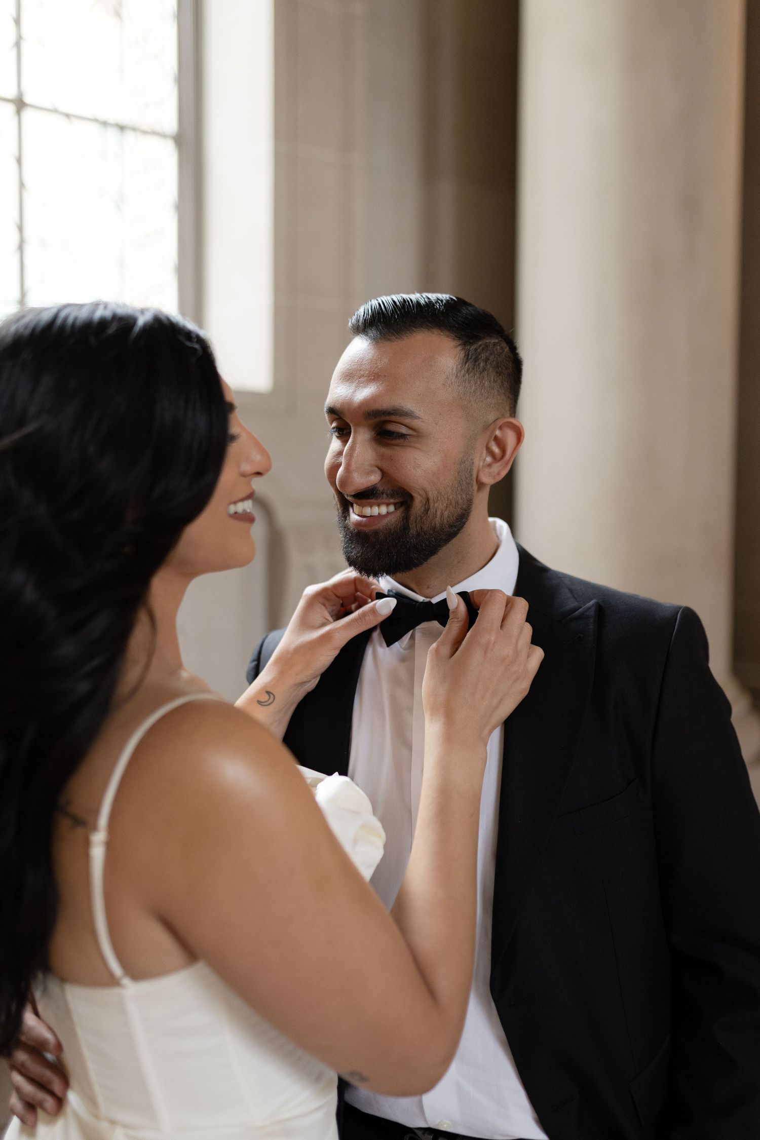 A bride in a white dress playfully touches her groom's beard during a romantic wedding photoshoot in a sunlit room.