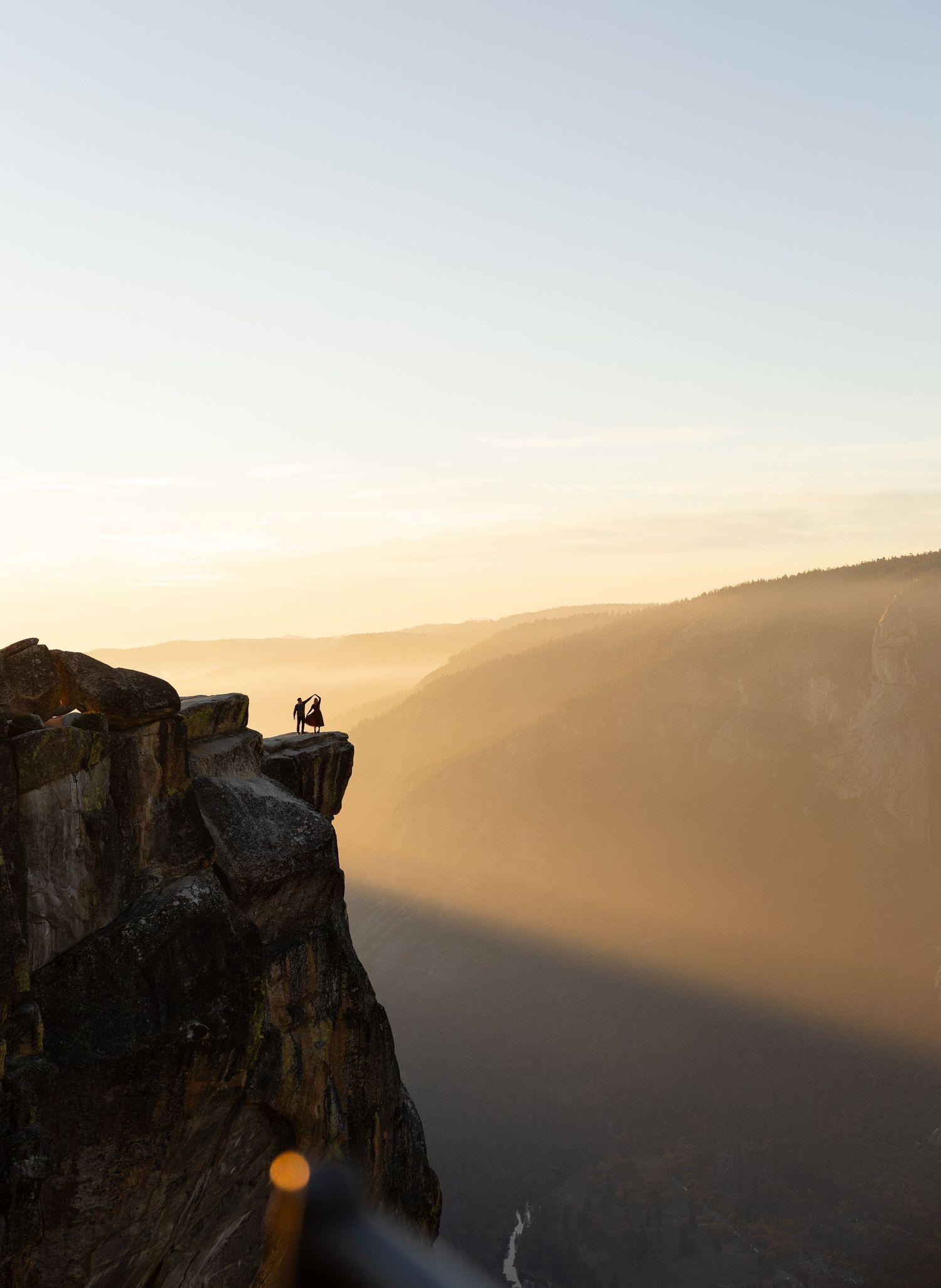 Distant figures stand on a towering cliff edge against a golden sunset landscape.