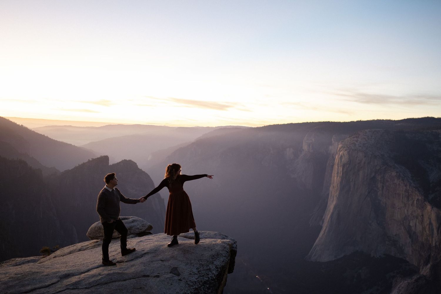 Two silhouettes stand on cliff edge at Yosemite's Taft Point during golden sunset over mountain valley.