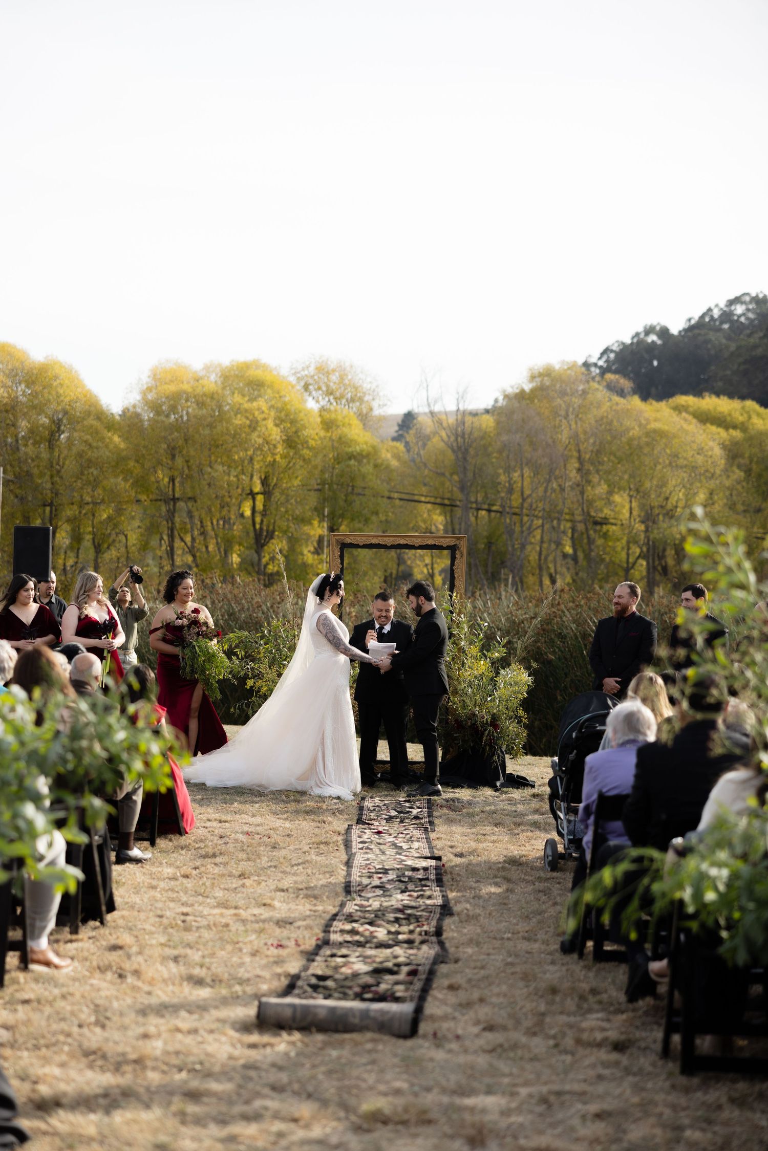 Wide view of outdoor wedding ceremony with wooden arch and guests seated on both sides of aisle.