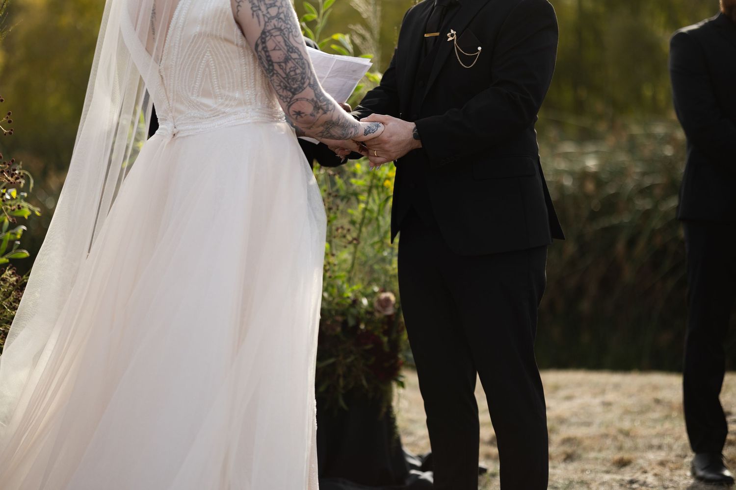 Close-up of joined hands during a wedding ceremony showing tattooed arms and white bridal gown details.