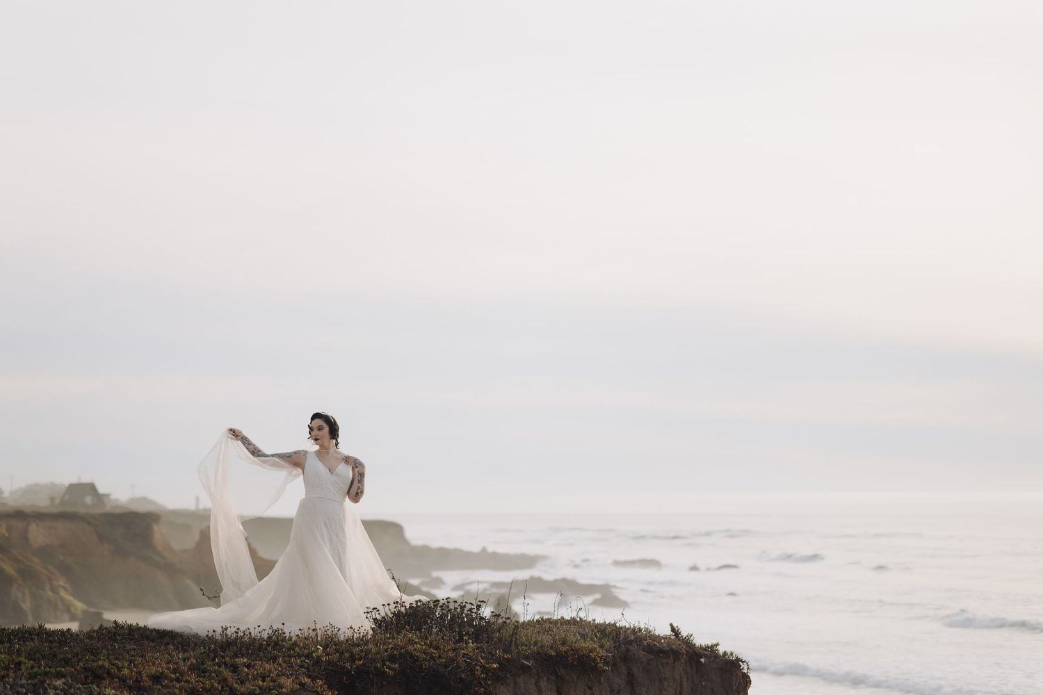 Bride in flowing white dress poses dramatically on coastal cliffs at sunset.