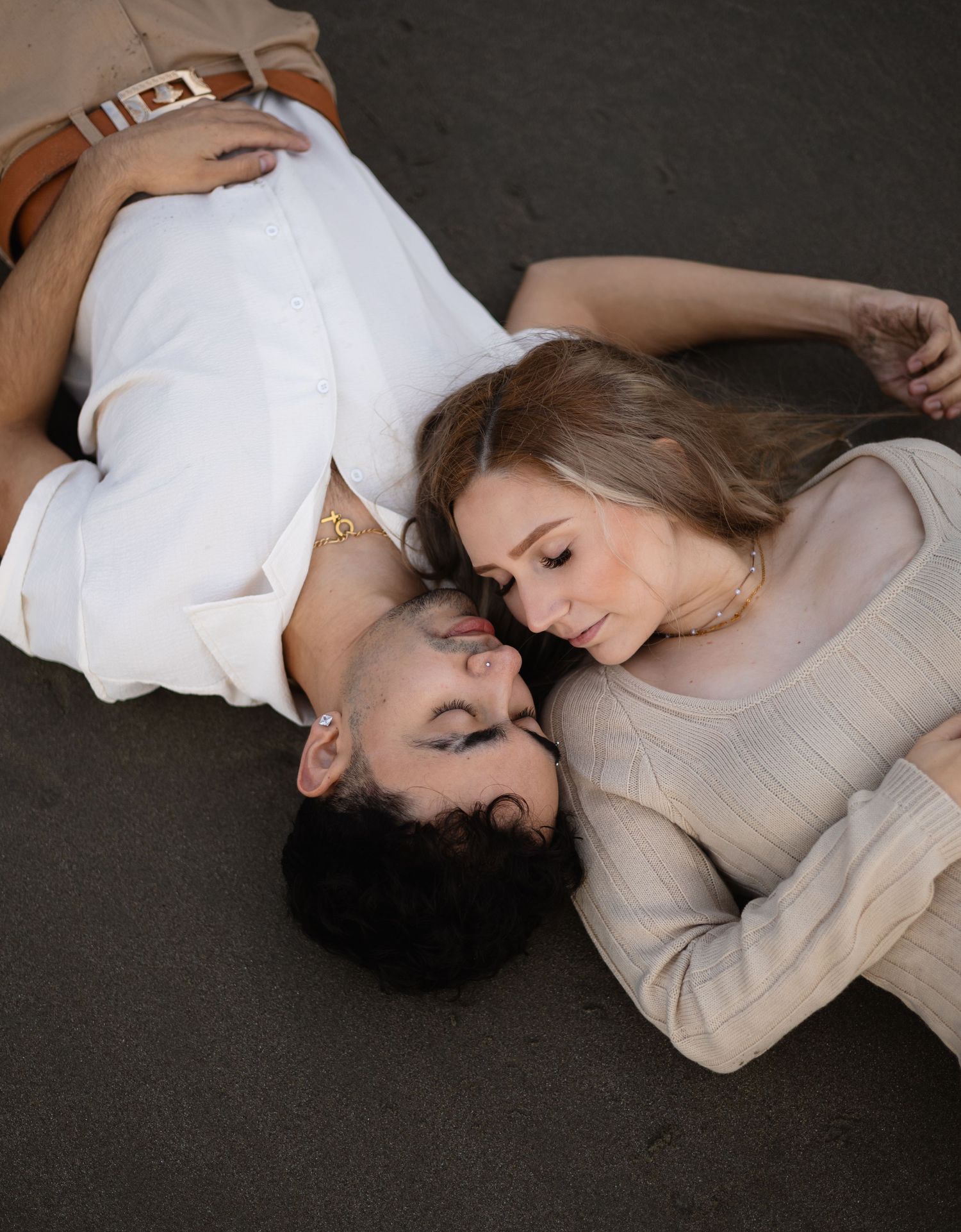 A couple lies close together on a dark surface in casual white and beige clothing.