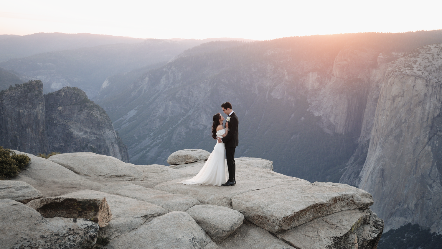 Bride and groom embracing on a rocky cliff edge at sunset in Yosemite National Park with dramatic valley views.