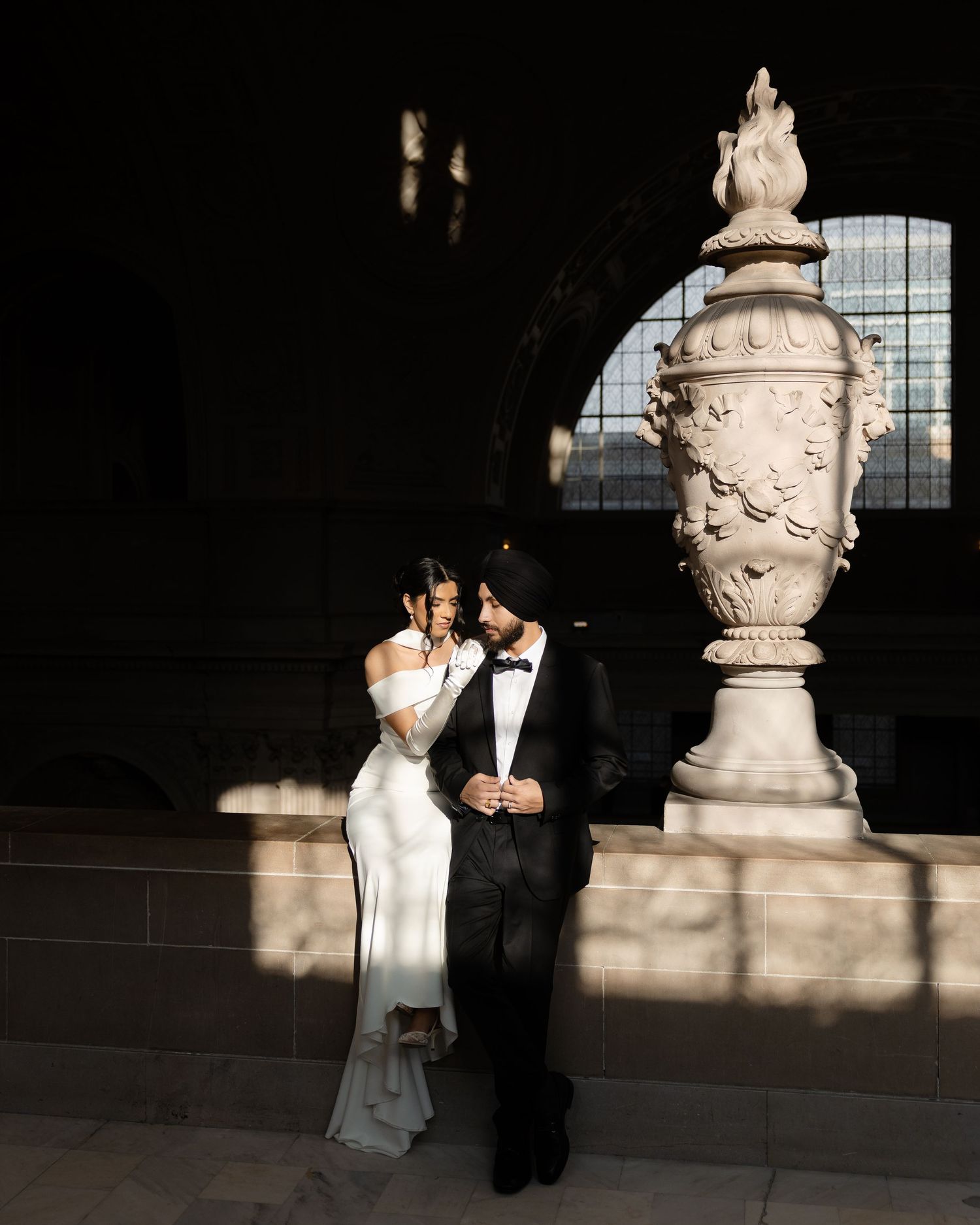 A couple in formal wedding attire stands in dramatic lighting against a stone wall with ornate architectural details.