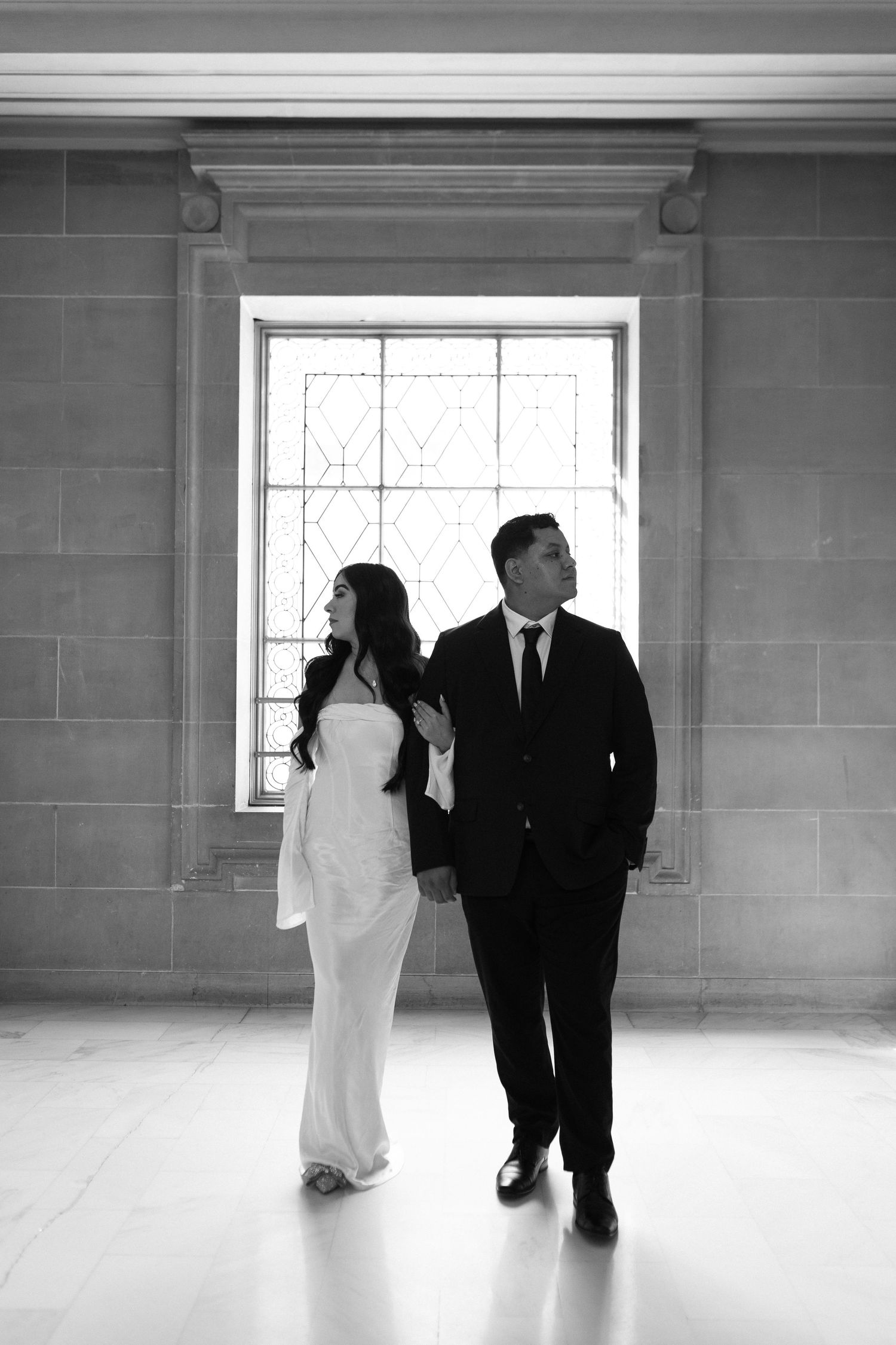 A black and white photo of newlyweds walking together in an elegant corridor with natural window light.