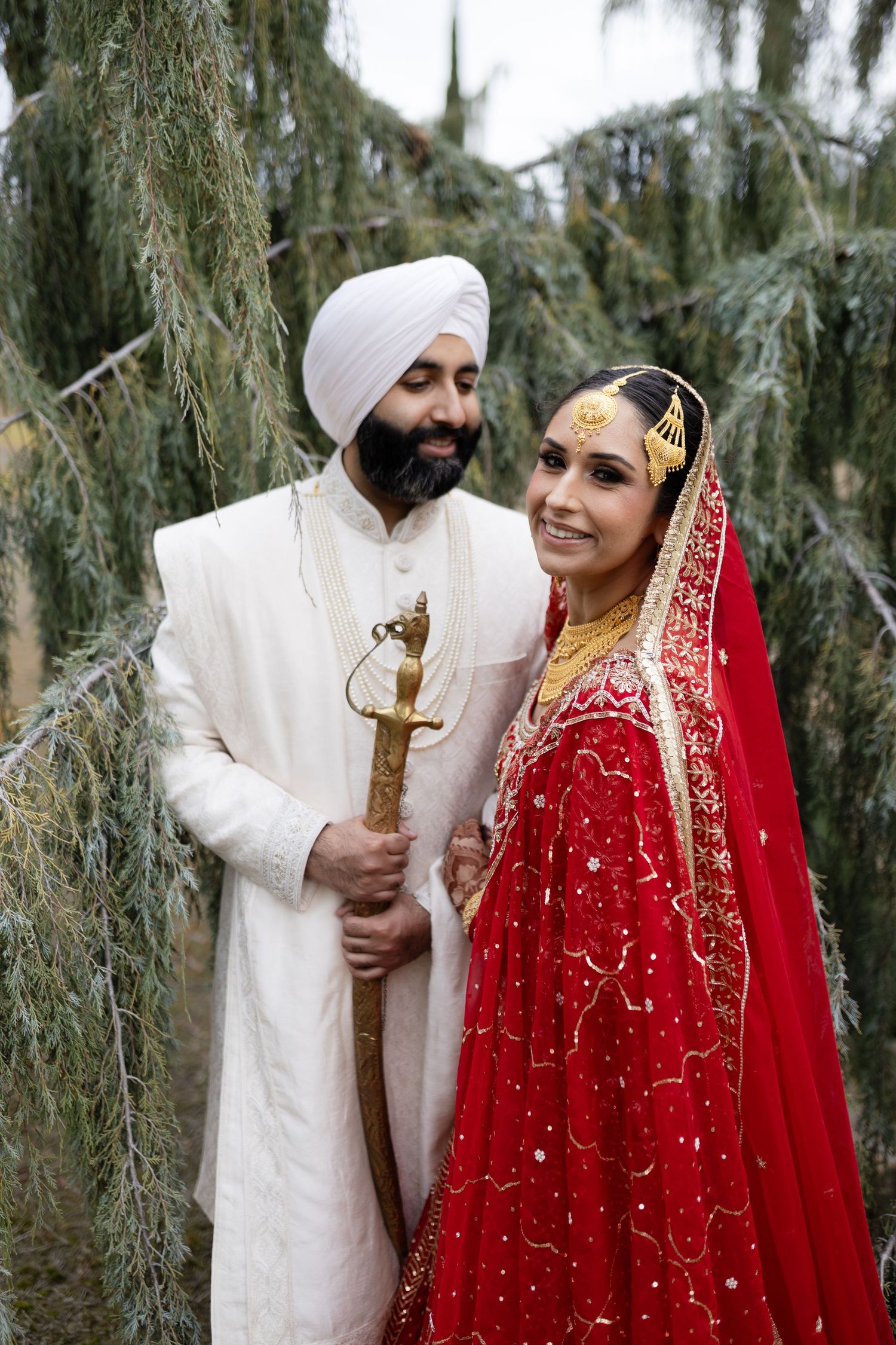 A traditional Sikh wedding couple poses together outdoors among trees, with the bride wearing a stunning red embroidered dress.