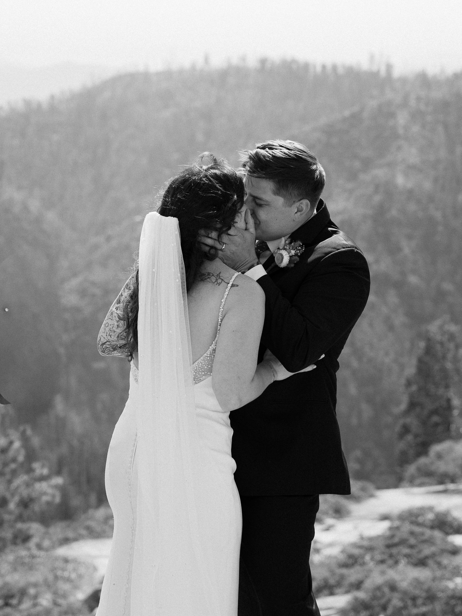 Black and white photo of a couple sharing a first kiss in wedding attire with misty mountain forest backdrop.