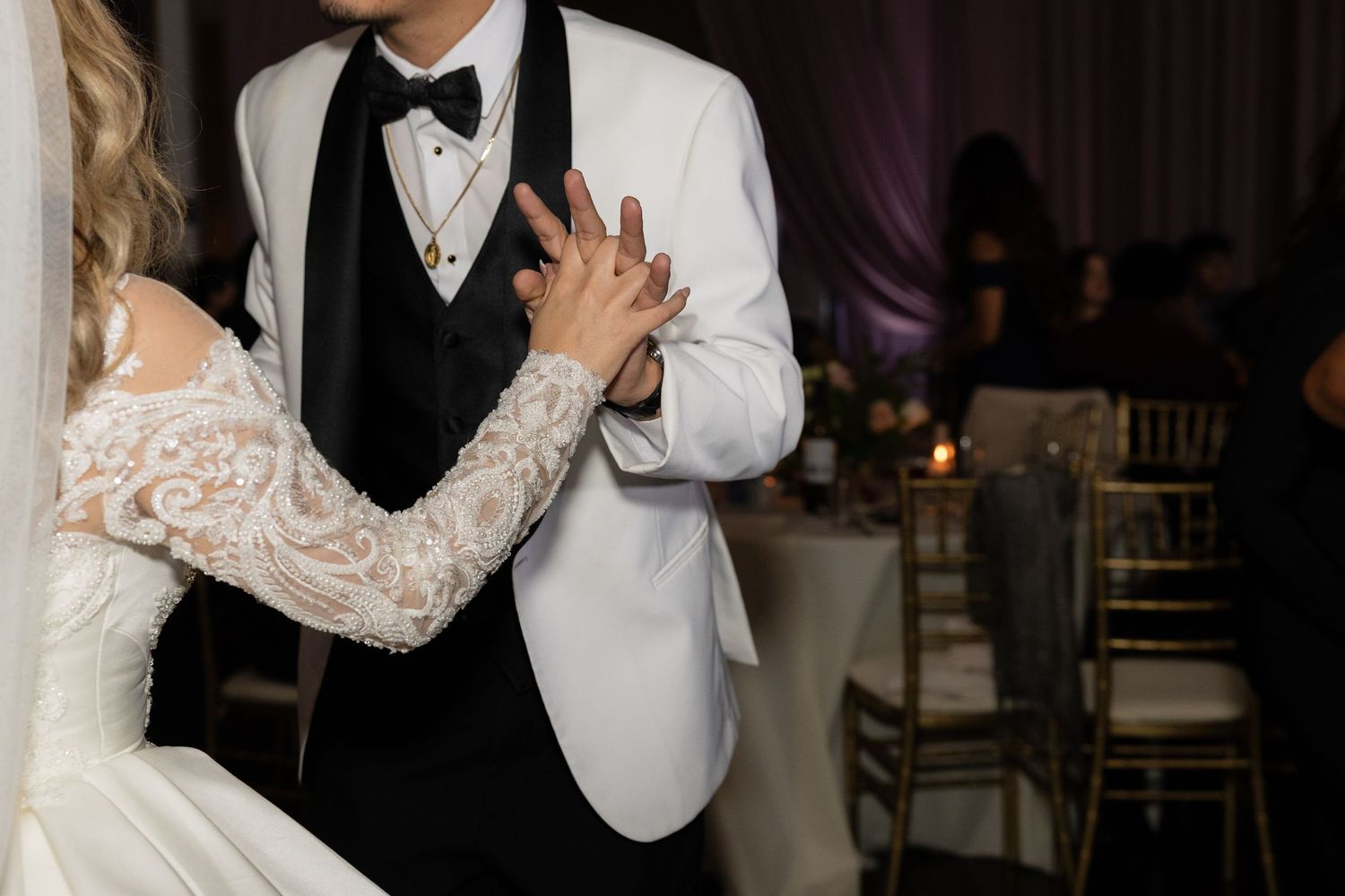 Close-up of couple's hands during wedding dance showing detailed lace sleeve and white tuxedo jacket.