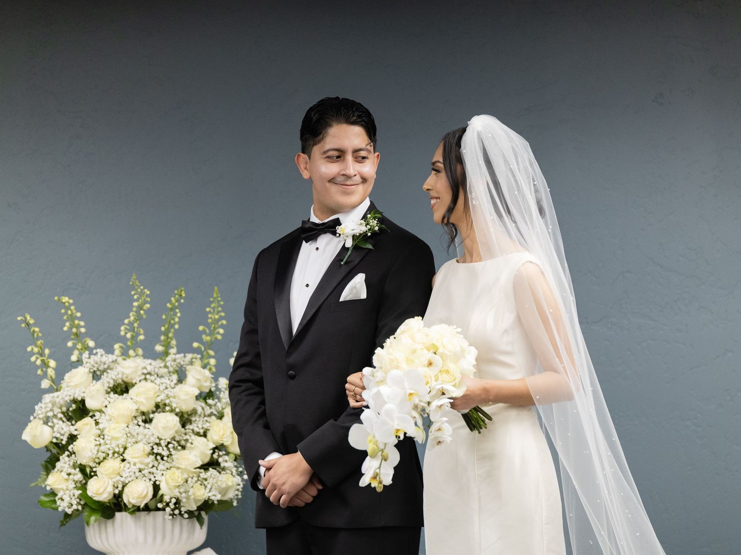 Wedding couple posing with white floral arrangements against grey studio background.