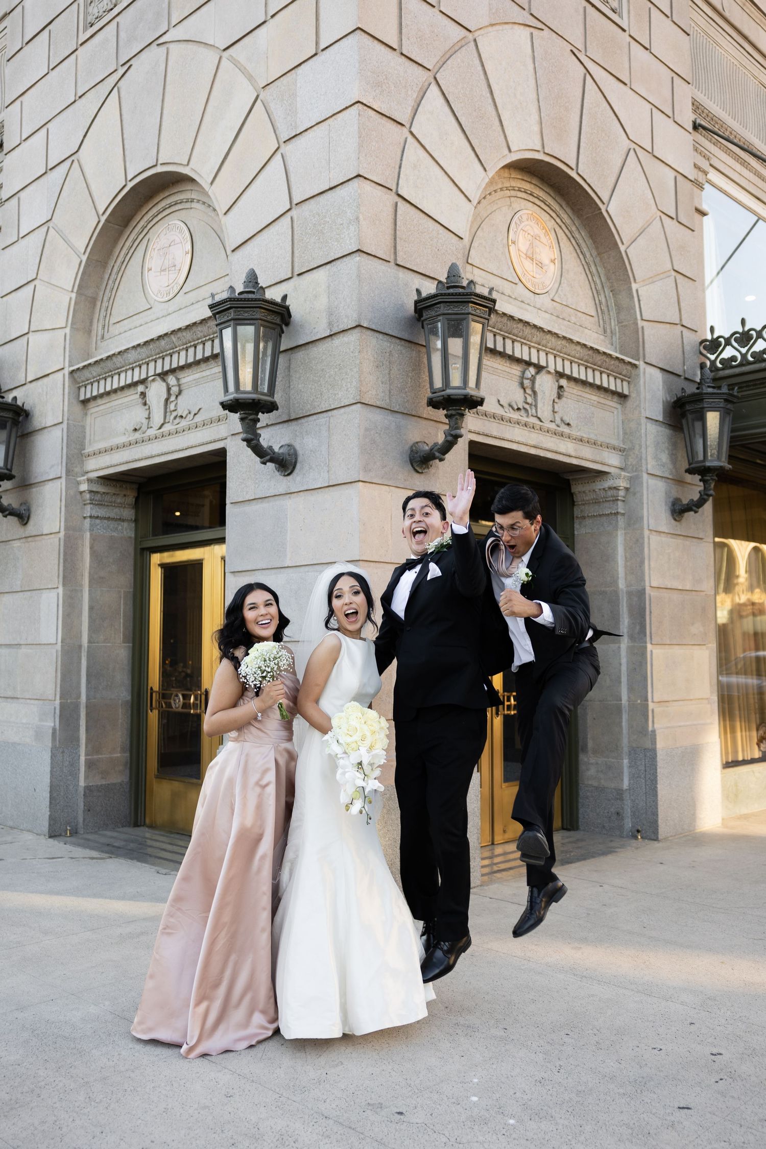 Wedding party members pose joyfully outside historic building with ornate lanterns.