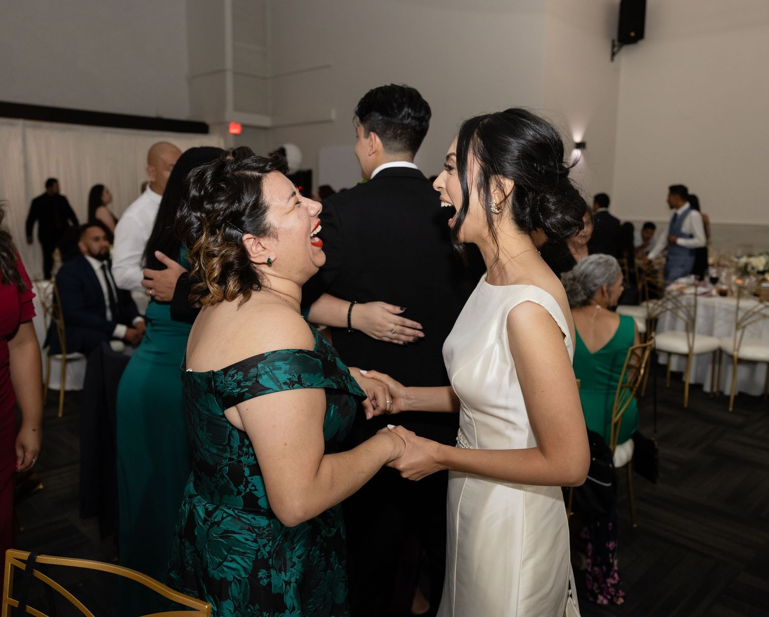 Wedding guests in emerald green and white dresses dancing together at an indoor reception celebration.