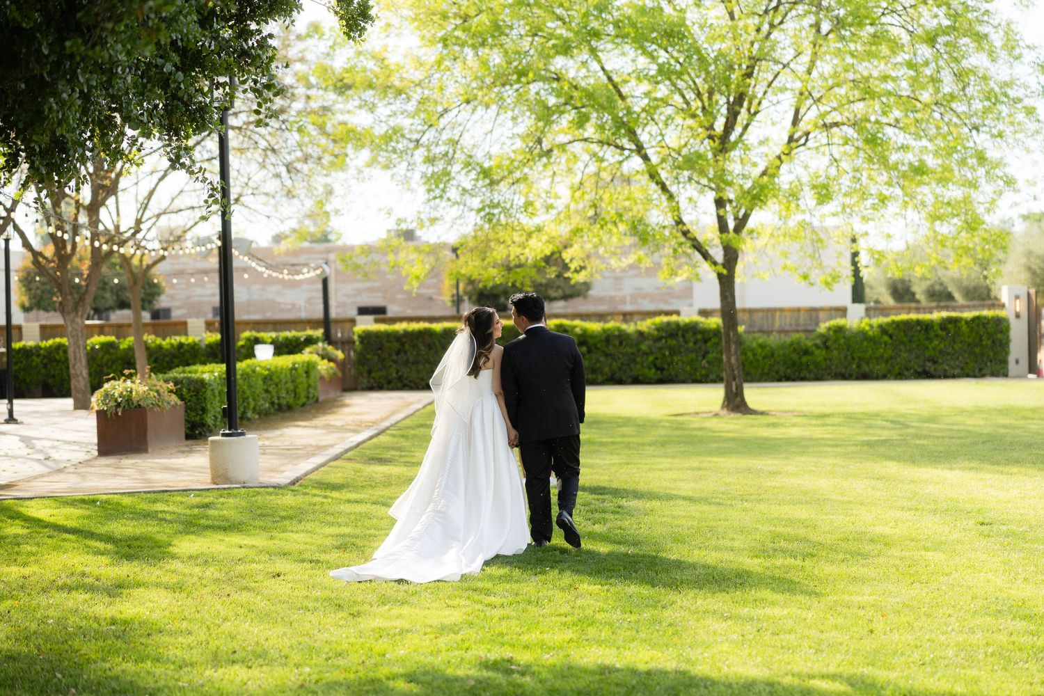Bride and groom walking hand in hand across lush green lawn surrounded by manicured hedges and trees at outdoor wedding.