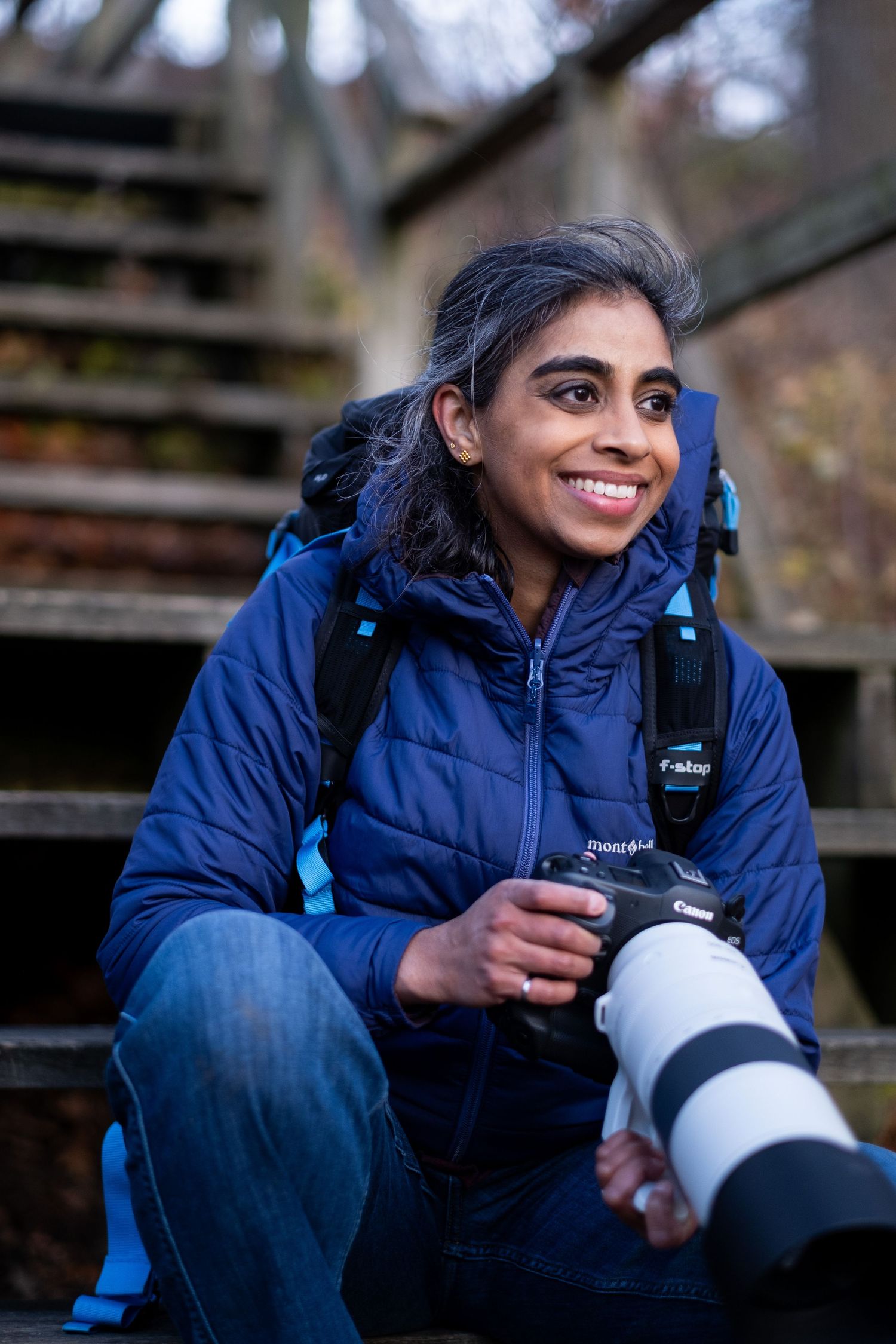 A south Asian woman wearing a blue jacket is sitting on wooden stairs and is holding a large camera.