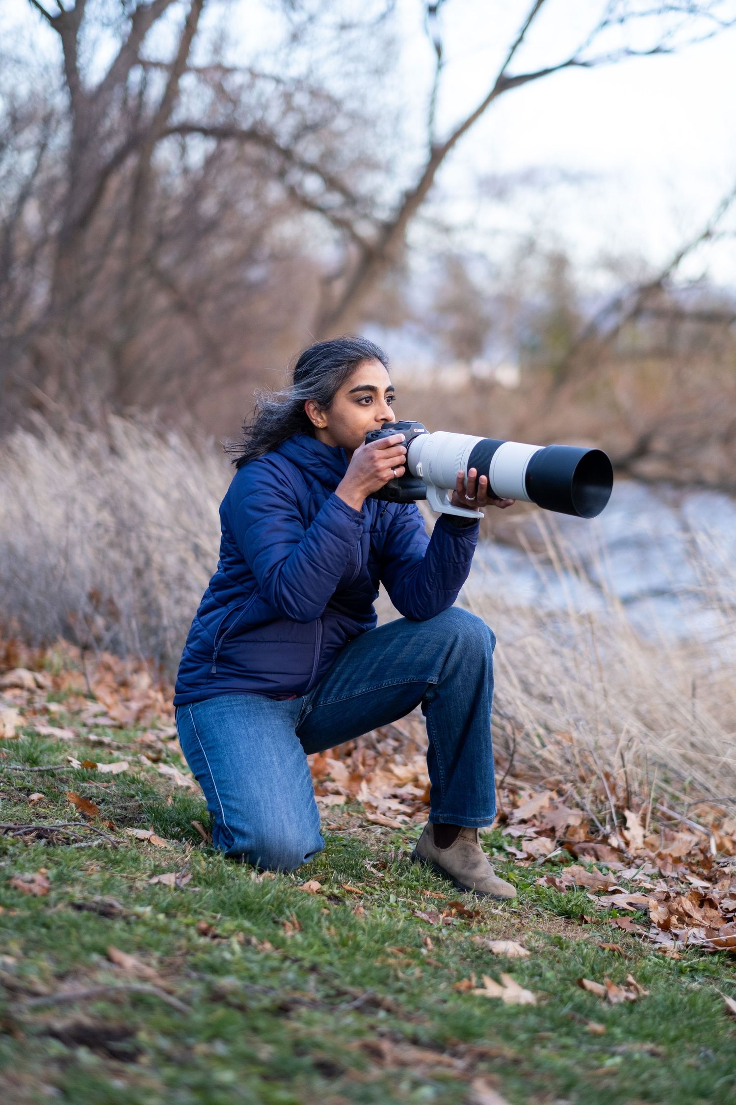 A south Asian woman wearing a blue jacket is kneeling on grass and is taking a photo with a large camera.