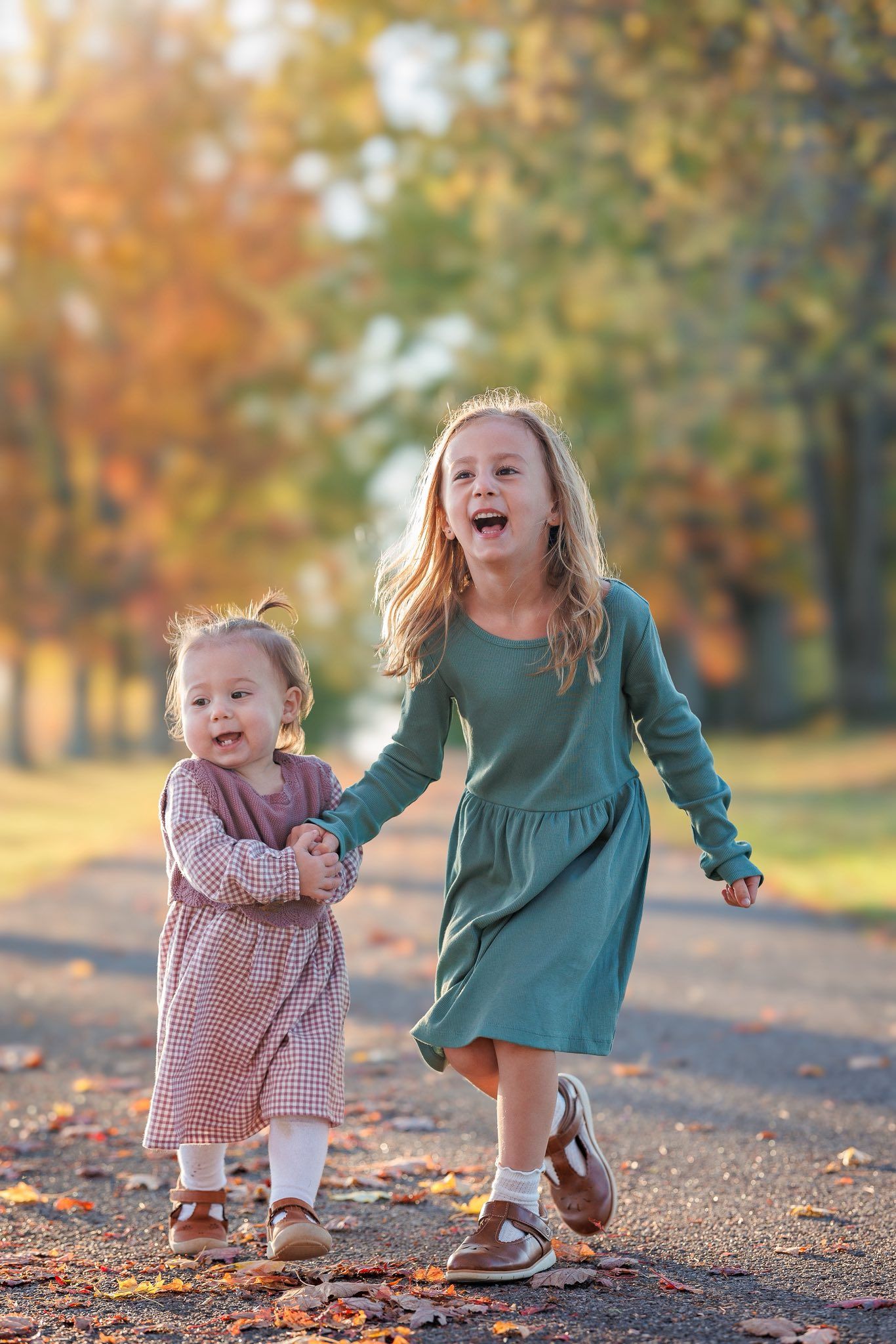 Two children laugh and hold hands while running through autumn leaves on a path.