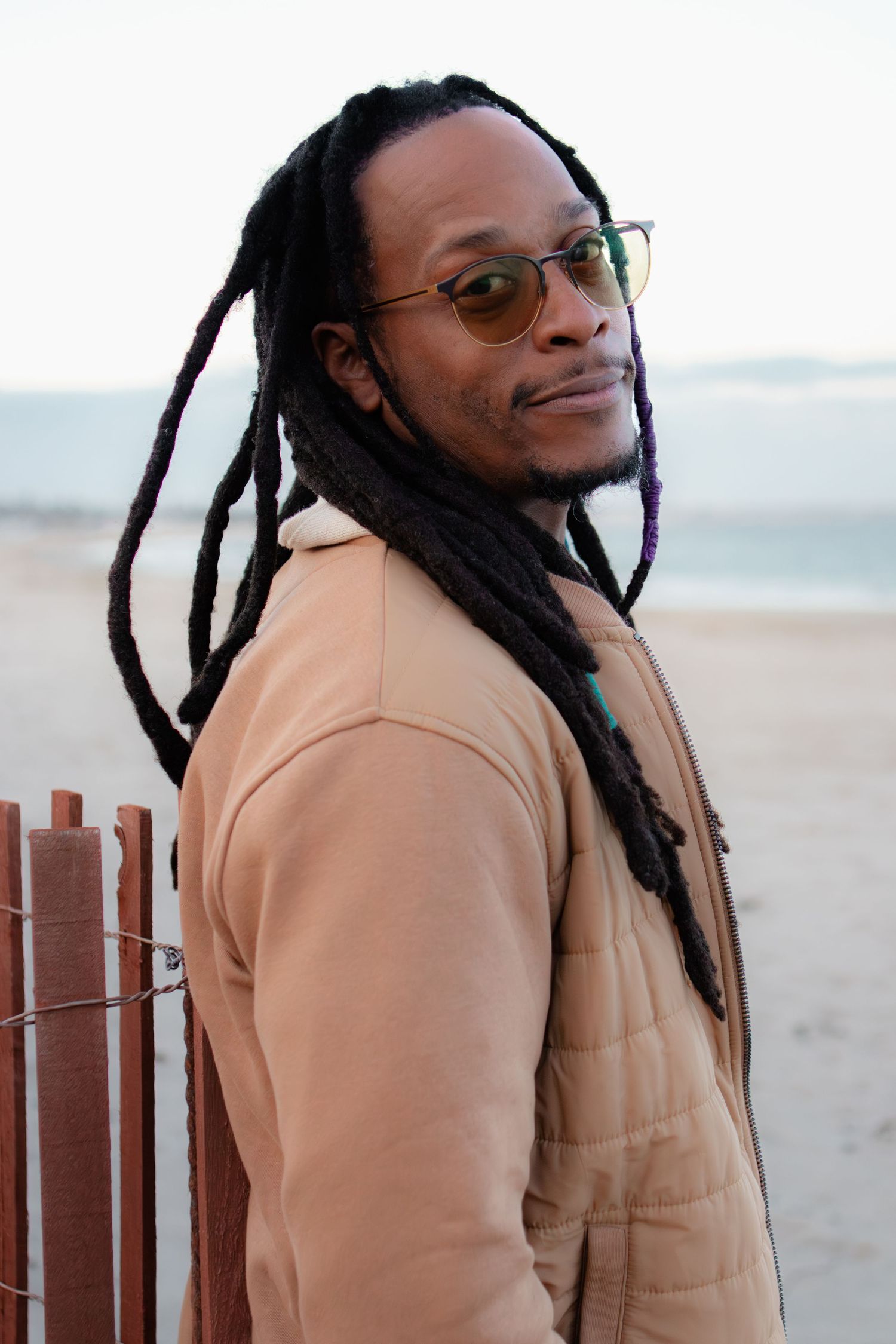 Young Black man with long dreadlocks and glasses wearing a beige coat, standing near a beach fence.
