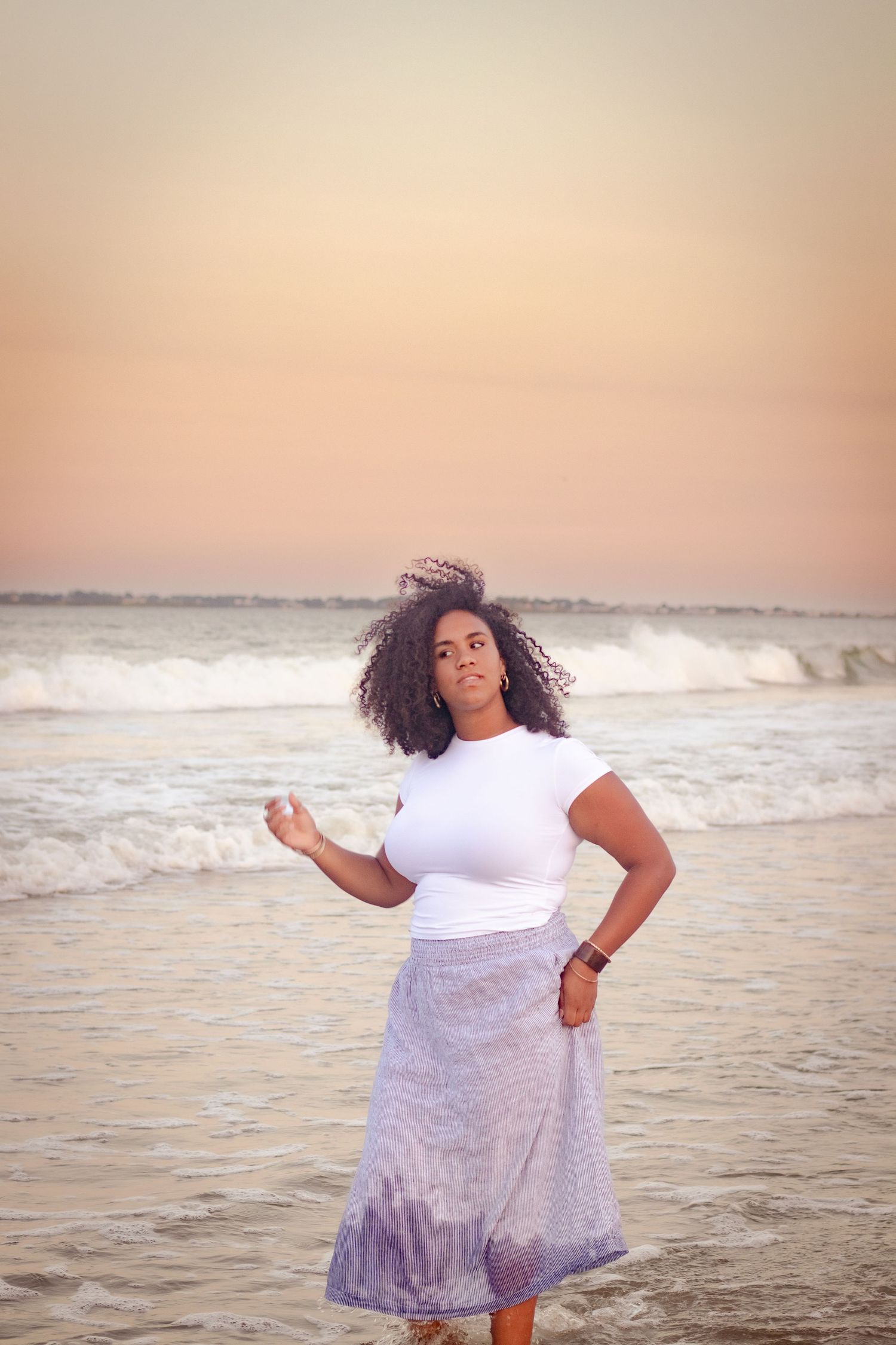 Person in white dress twirling on beach at sunset with ocean waves in background.