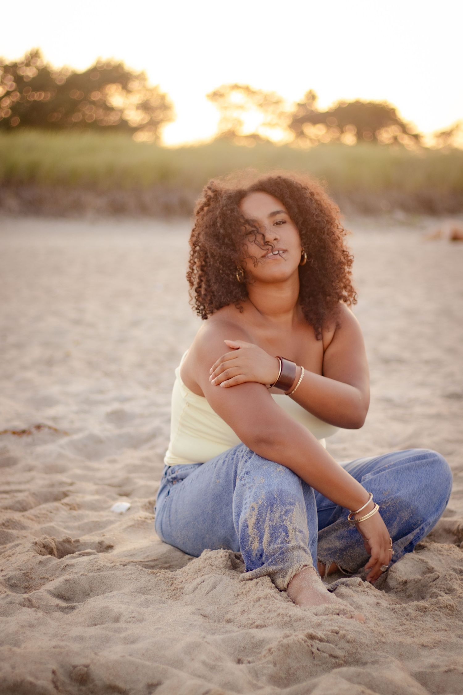 Person sitting casually on sandy beach at sunset wearing jeans.