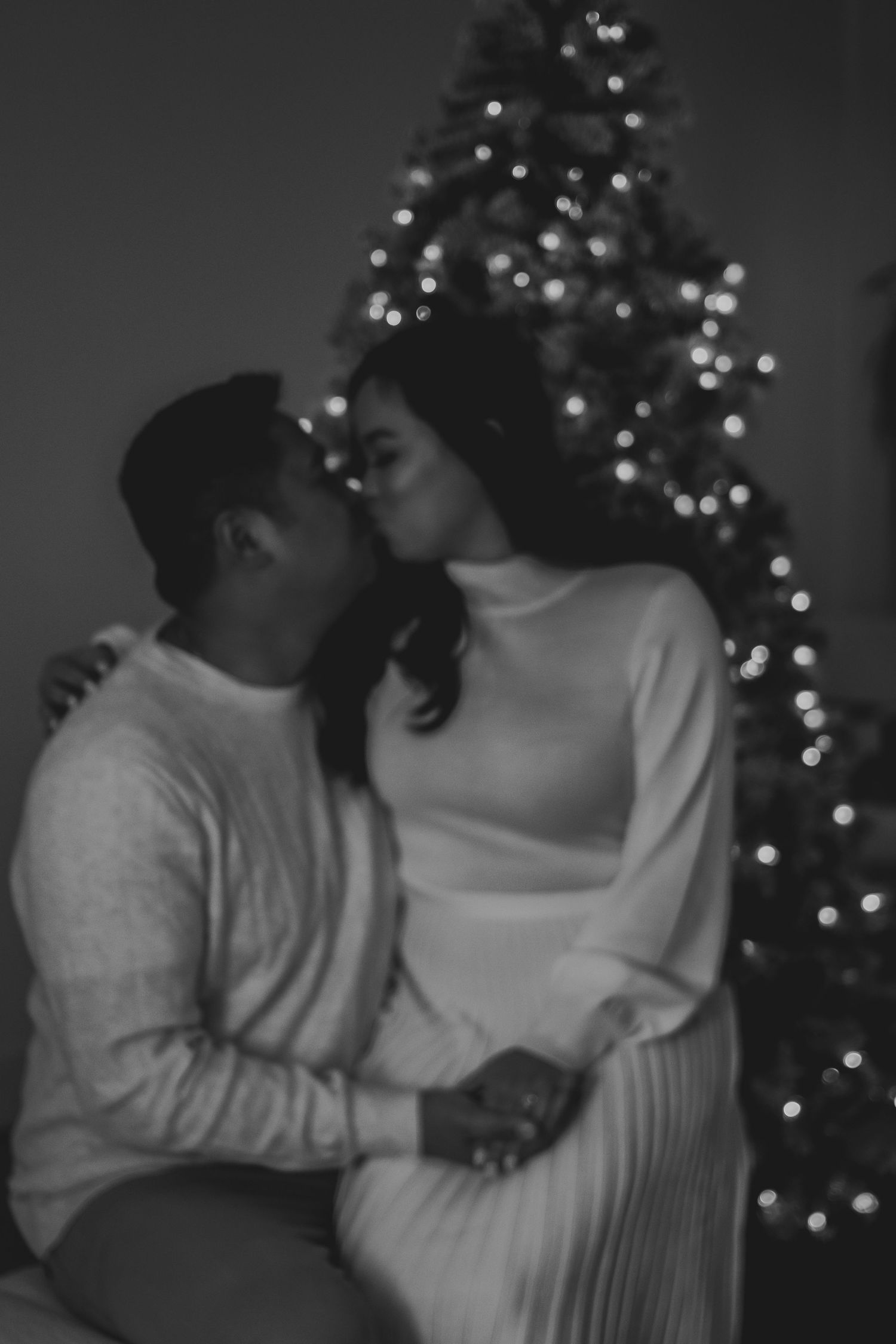 A romantic couple shares an intimate moment in front of a Christmas tree with twinkling lights in black and white.