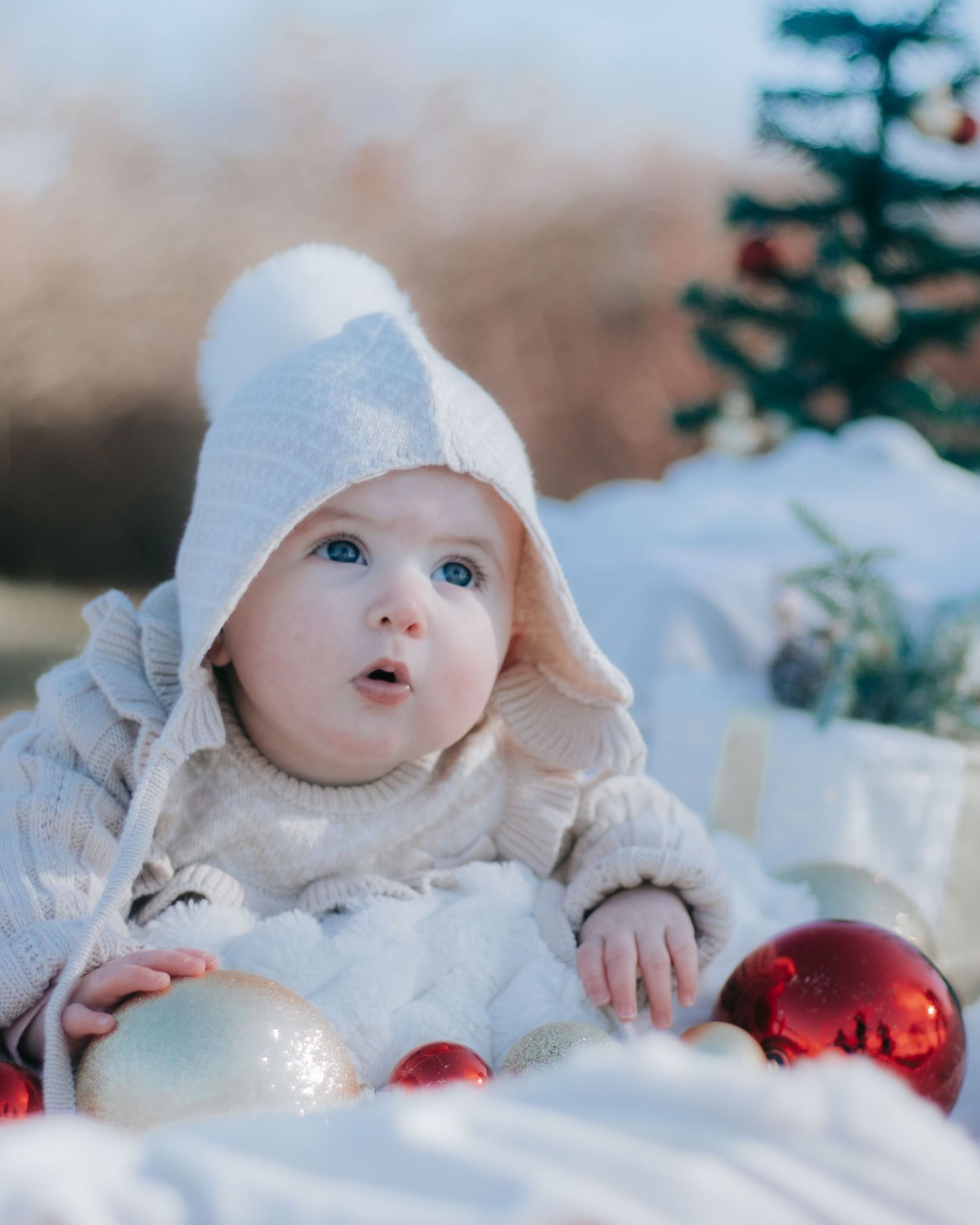 Baby in white winter clothing lies in snow with red Christmas ornaments.