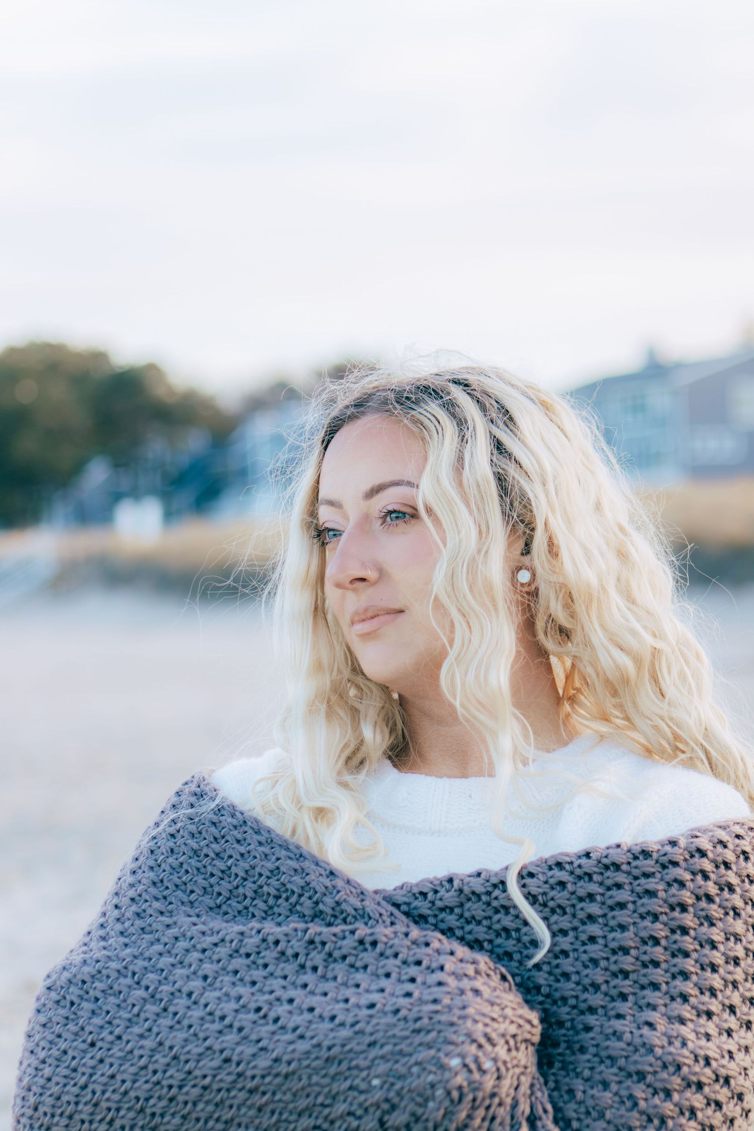 Person with wavy blonde hair wrapped in a cozy gray knit blanket on a cloudy day outdoors.