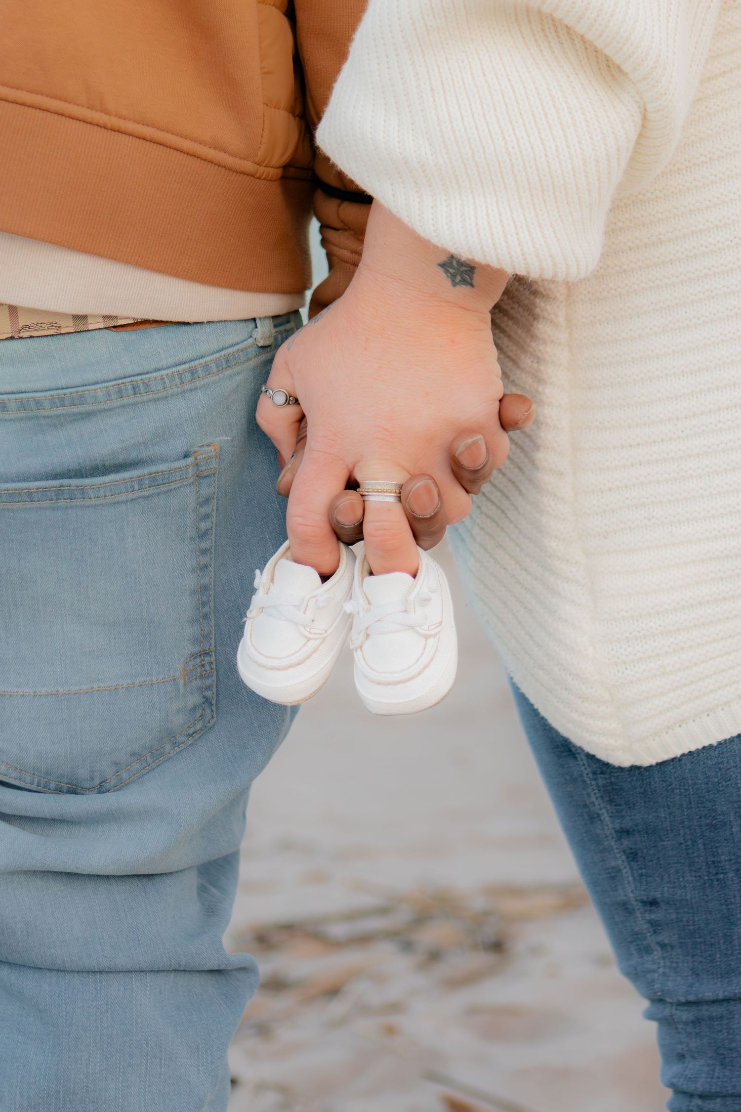 Couple holding hands and tiny white baby sneakers on a sandy beach, announcing pregnancy.